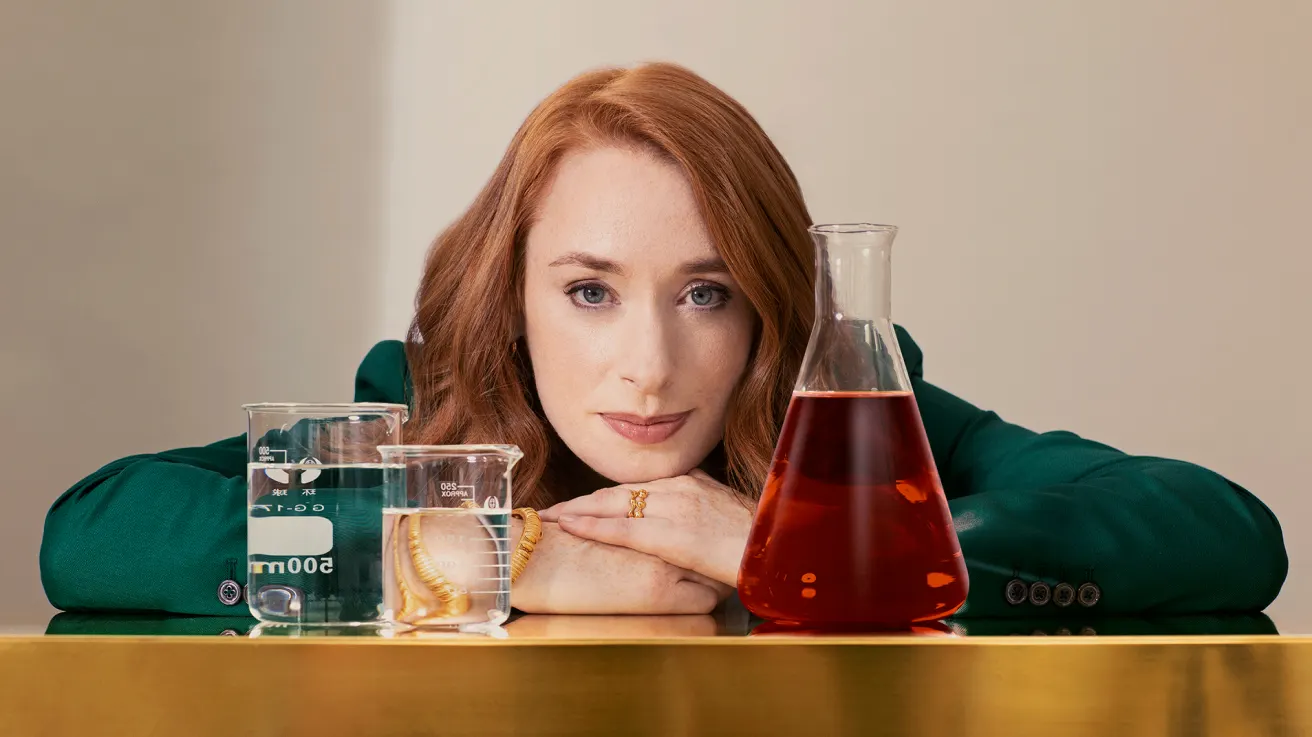 A woman with long red hair and a green blazer rests her chin on her hands, surrounded by three glass containers: two beakers with clear liquid and a worm, and a flask with a red liquid, on a reflective surface.