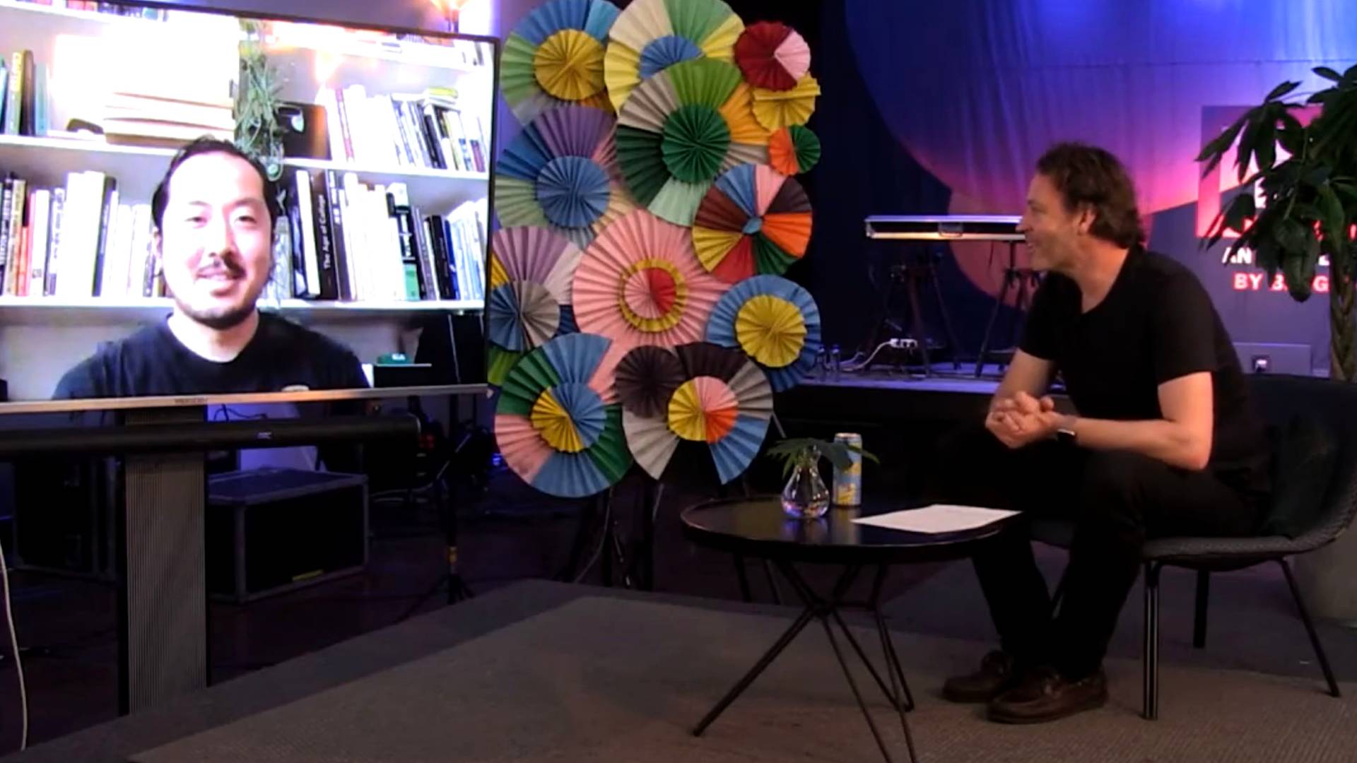 A man sits on stage at a small table, smiling and looking at a screen showing another man on a video call. Colorful paper fans decorate the background. The atmosphere is casual and relaxed.