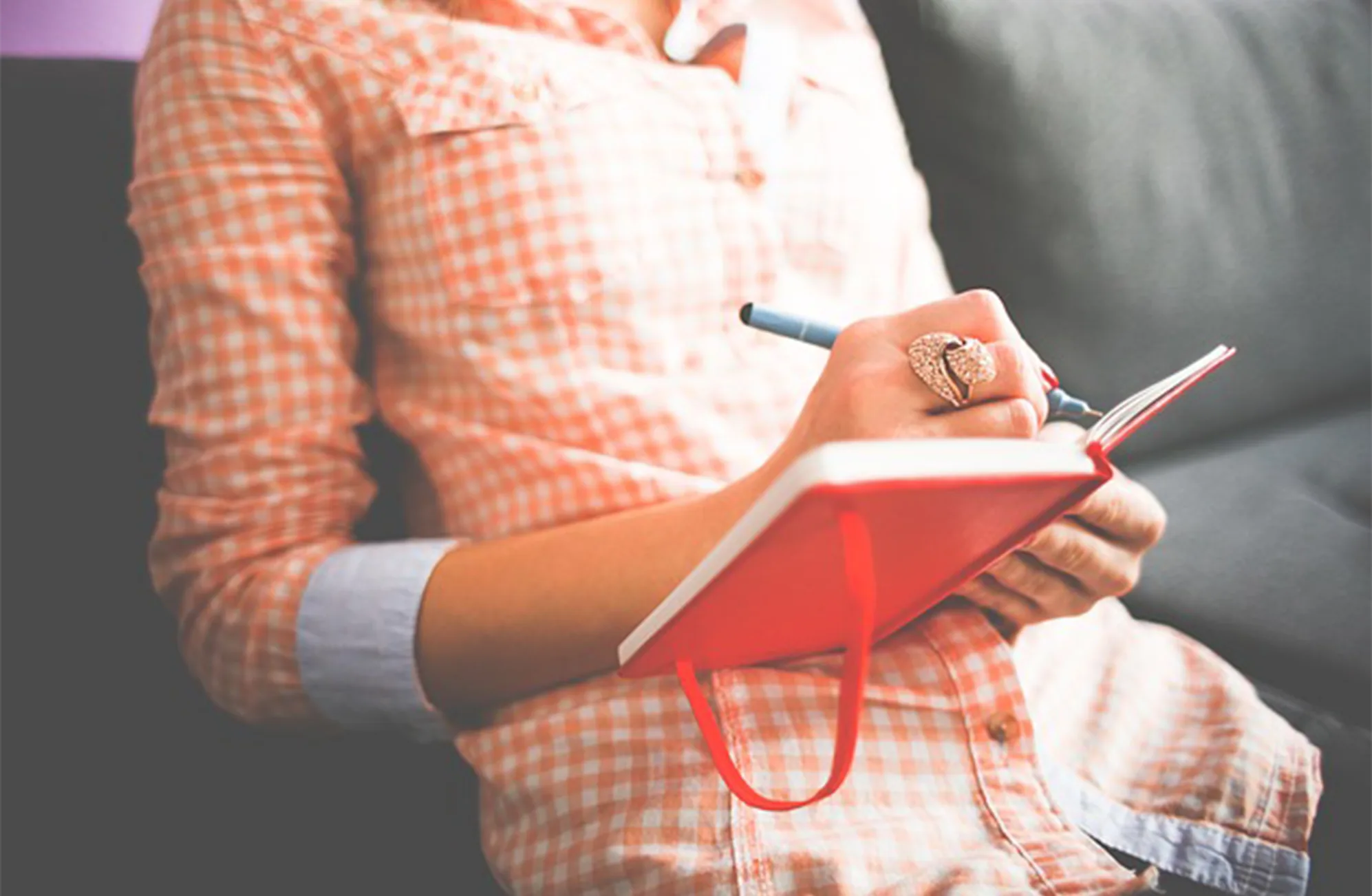 A person in a peach plaid shirt sits on a couch, holding a red notebook and writing with a pen. The focus is on their hands and the notebook, and their face is not visible.