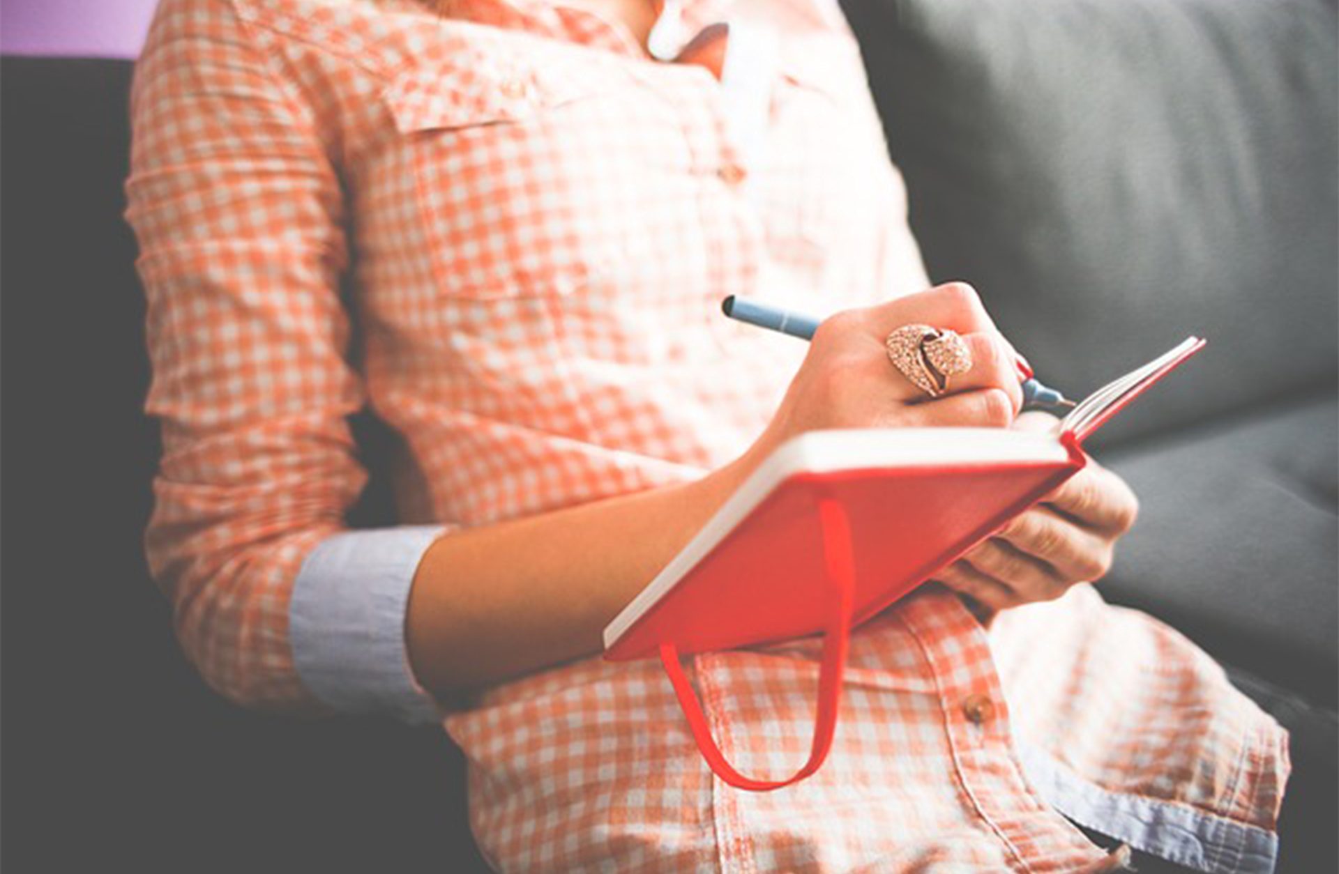A person in a peach plaid shirt sits on a couch, holding a red notebook and writing with a pen. The focus is on their hands and the notebook, and their face is not visible.