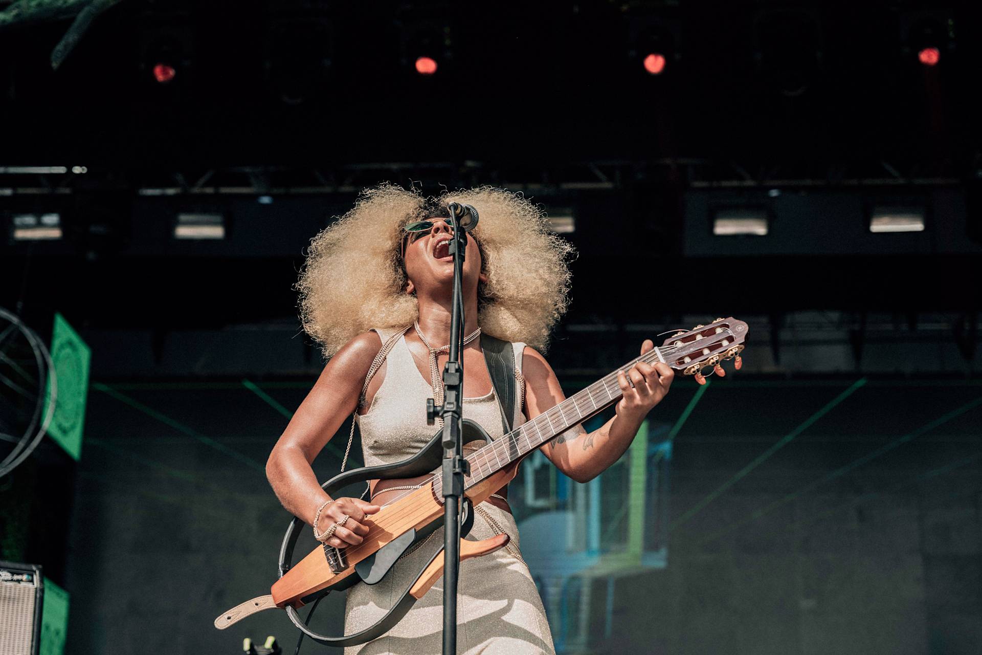 A musician with curly blond hair and sunglasses passionately sings and plays an acoustic guitar on stage, under colorful stage lights, during a live outdoor performance.