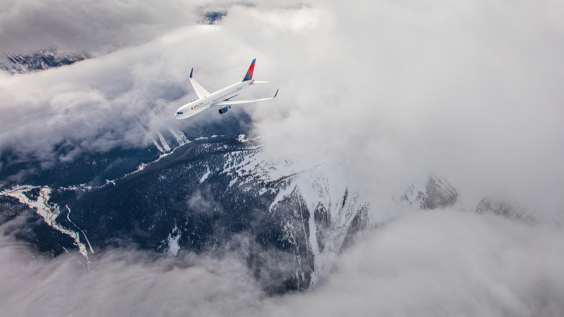 A commercial airplane flies above snow-covered mountains partly obscured by clouds, with a winding river visible below.