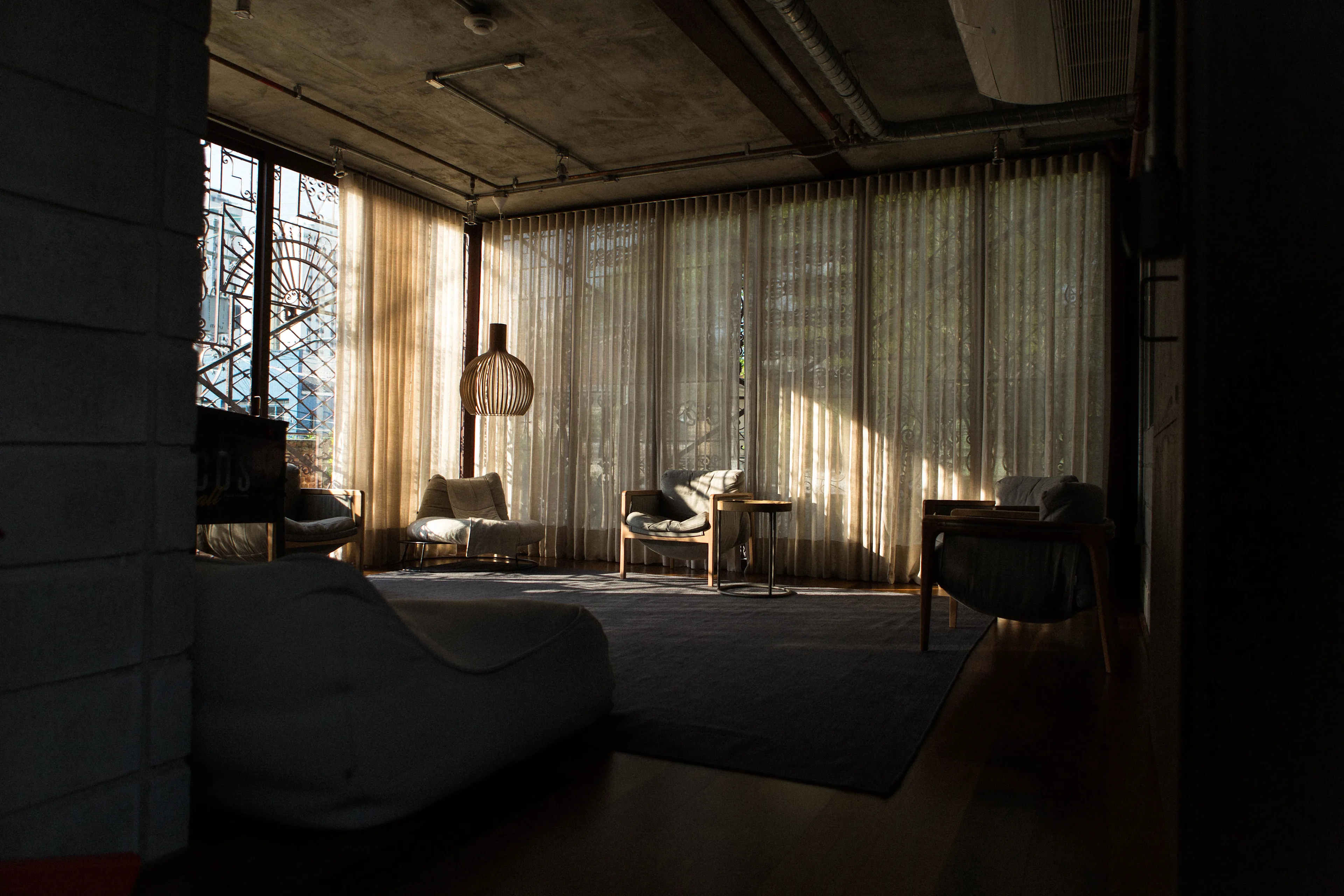 A modern living room with large floor-to-ceiling windows covered by sheer curtains, letting in soft sunlight. The room has armchairs, a small table, and a hanging lamp, with cozy, minimalist decor.