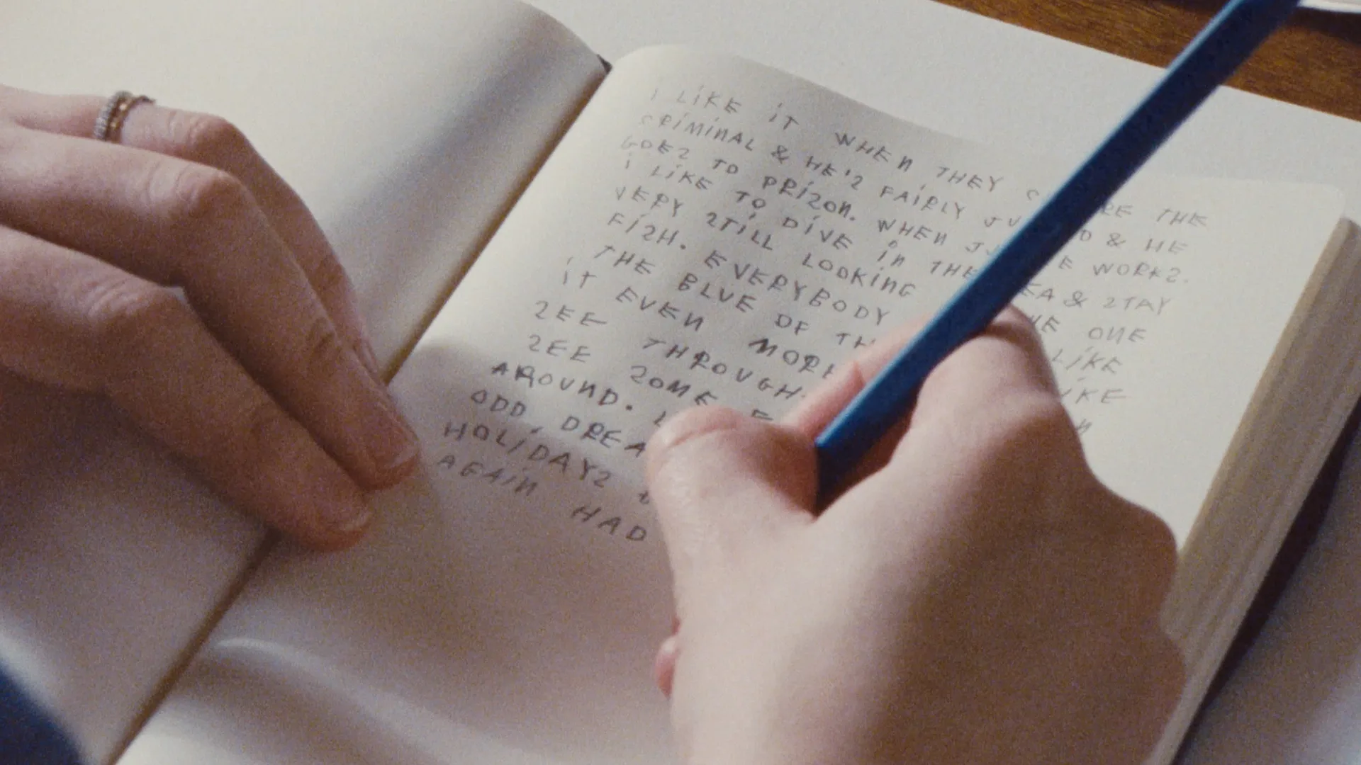 A person writes in a notebook with a blue pencil. The handwritten text is in all capital letters and fills most of the page. Only the persons hands and part of the notebook are visible.