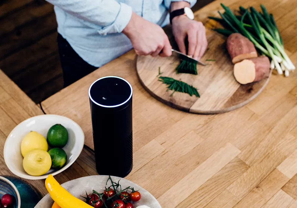 A person chops green onions on a wooden cutting board next to a smart speaker, with bowls of fruits and vegetables, including lemons, limes, cherry tomatoes, and a banana, on a wooden kitchen counter.