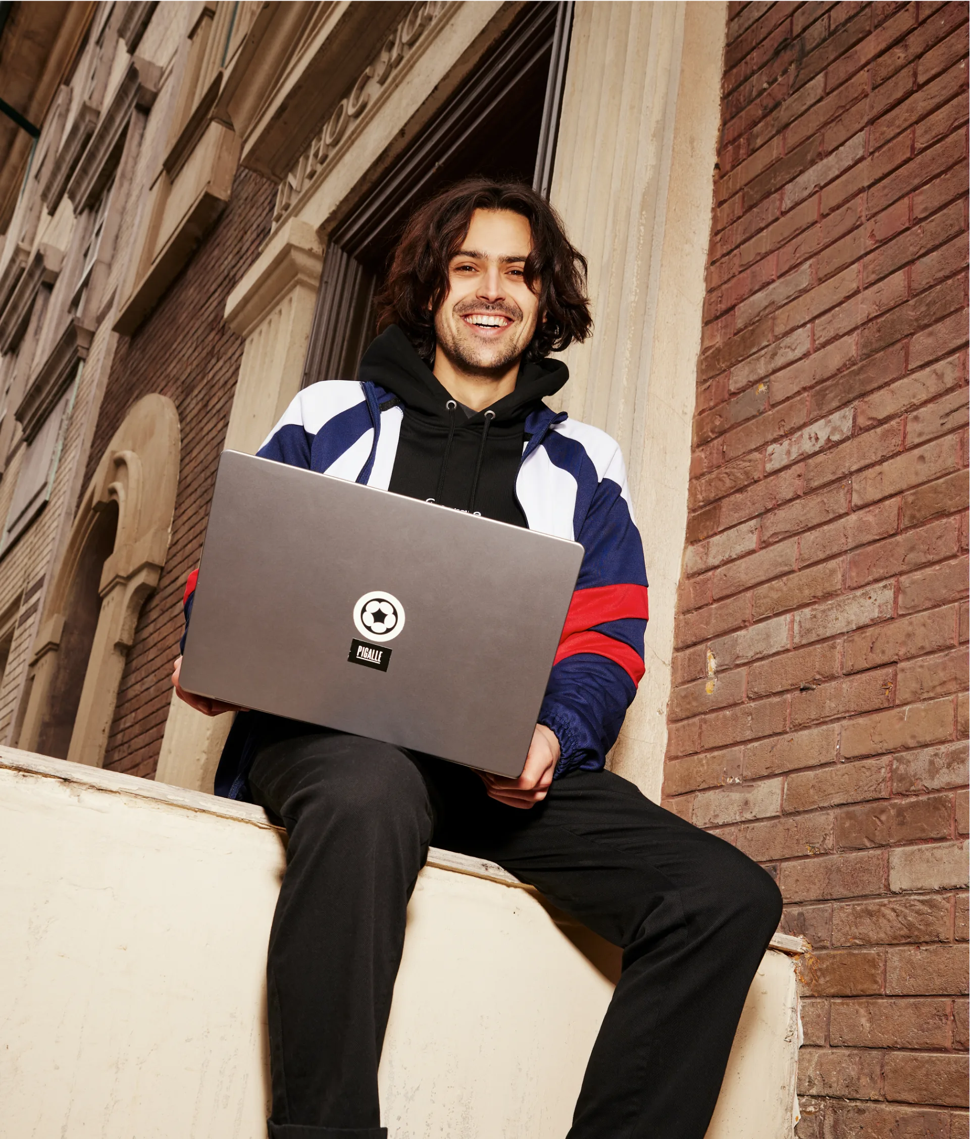A smiling person with shoulder-length hair sits on a ledge, holding a laptop, against a brick building backdrop. They wear a black hoodie and a blue, white, and red jacket.