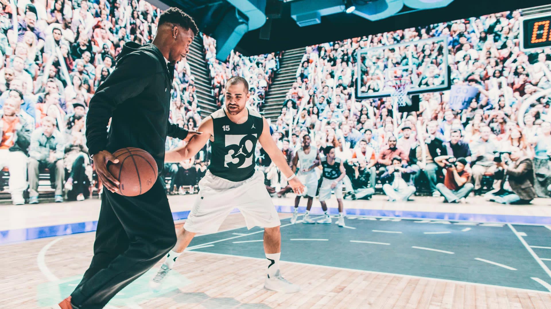 Two basketball players are in action on the court; one is dribbling the ball while the other defends. A large, cheering crowd fills the stands in the brightly lit arena.