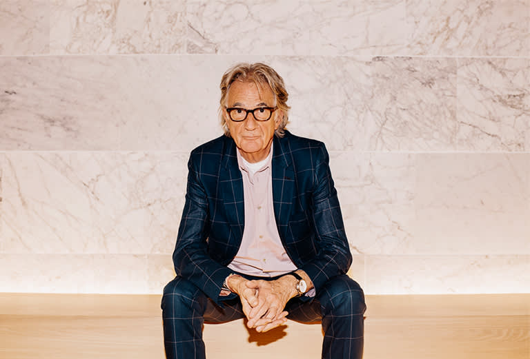 An older man with gray hair and glasses, wearing a blue plaid suit and light pink shirt, sits on a light wooden bench in front of a marble wall, looking directly at the camera with his hands clasped.