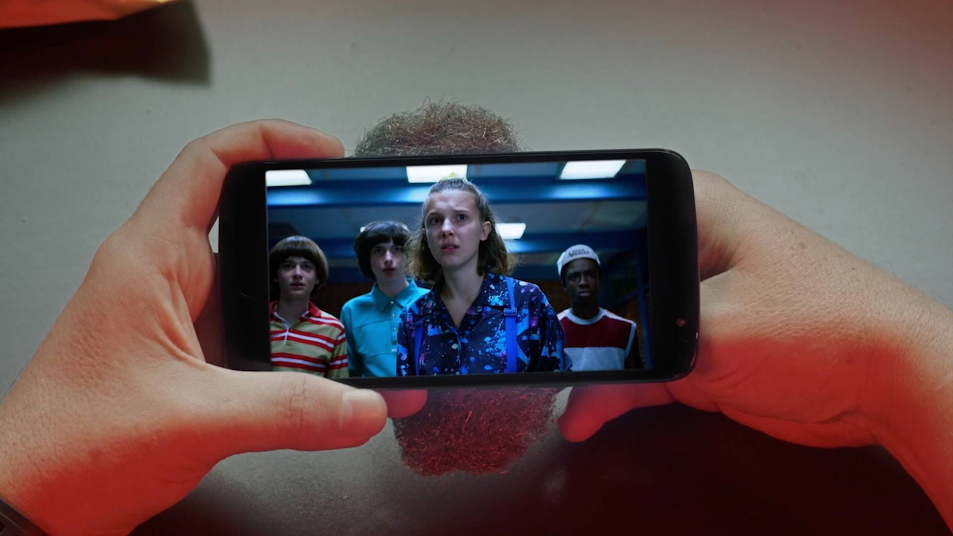 A person holds a smartphone displaying a scene from a TV show, where four teenagers stand together inside a building, looking ahead with serious expressions. The phone is held over a gray surface.