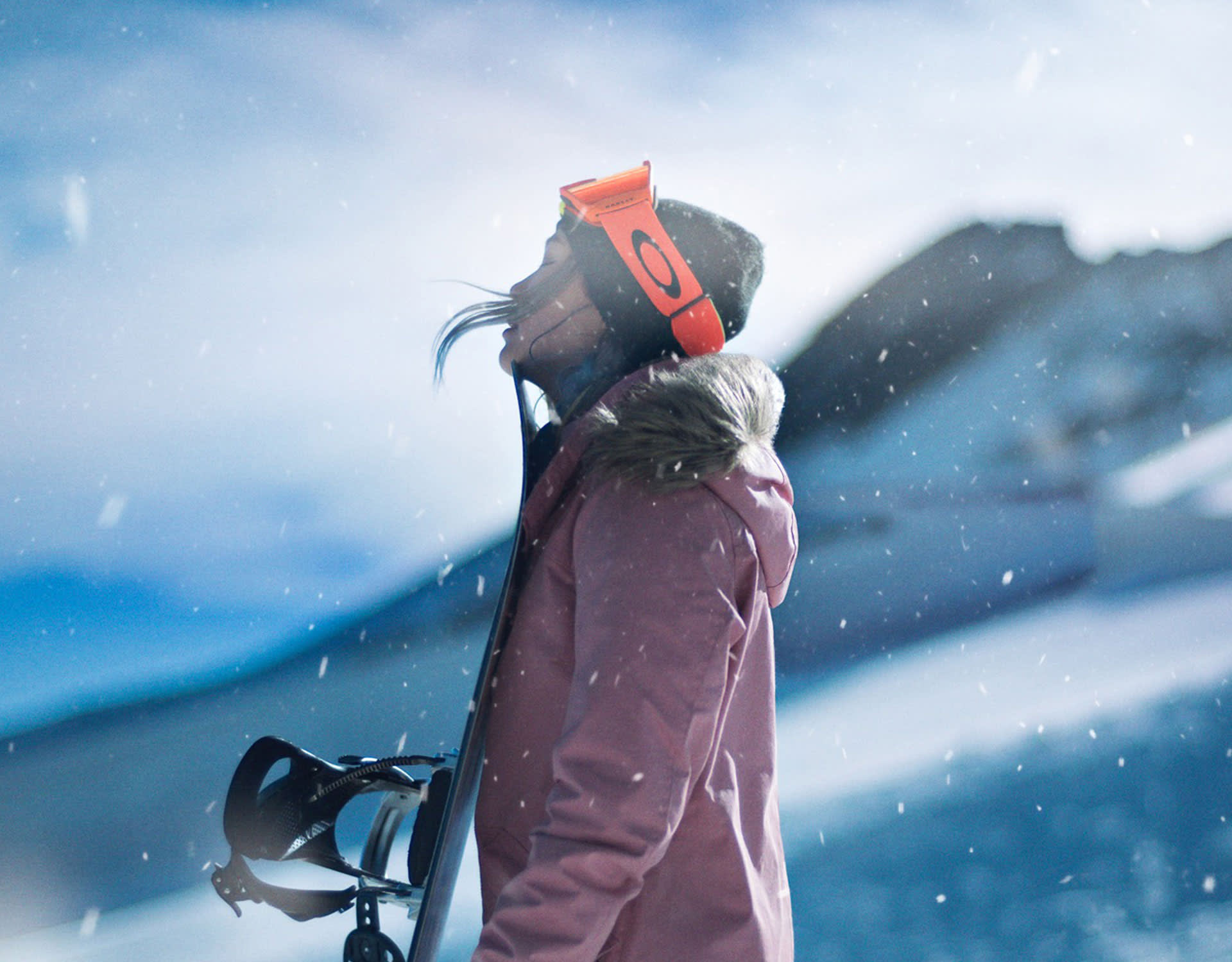 A person in a pink winter jacket and orange ski goggles stands in falling snow, holding skis over their shoulder, with a snowy mountain and blue sky in the background.