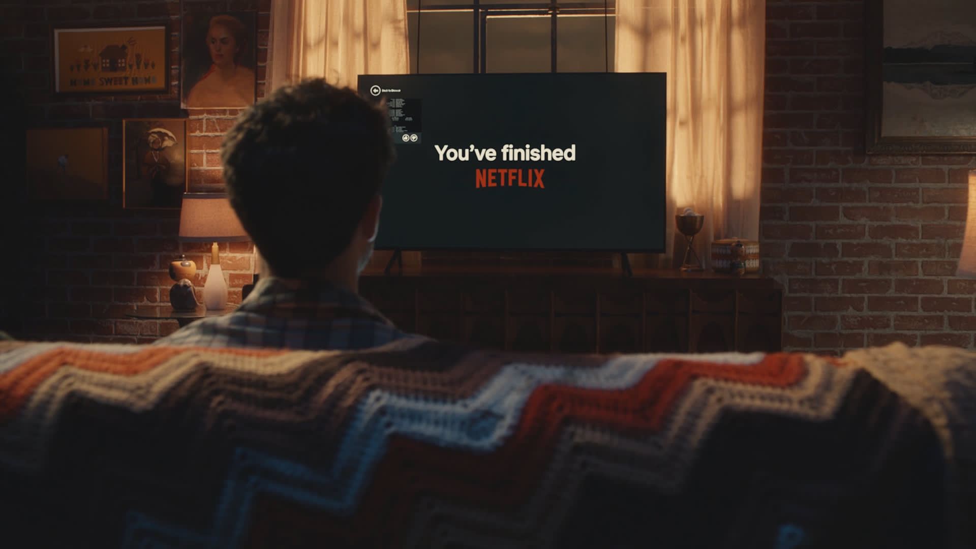 A person sits on a couch with a crocheted blanket, facing a TV displaying the message Youve finished NETFLIX in a cozy, dimly lit living room with curtains and a lamp.