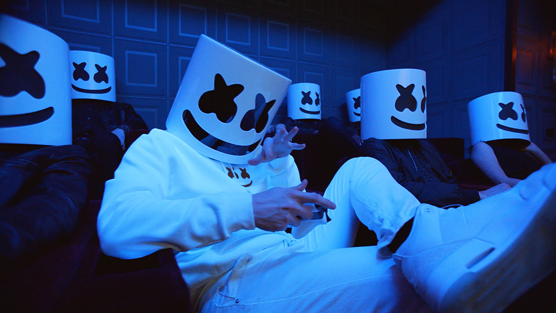 A group of people sit in a dimly lit room, all wearing white bucket-shaped masks with black cartoonish eyes and smiles, resembling the Marshmello DJ helmet. The lighting casts a blue hue over the scene.