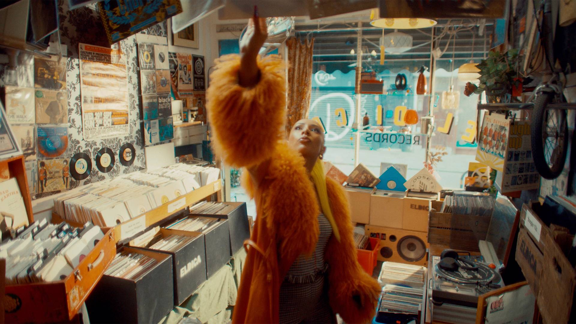 Eliza Rose in a bright orange fur coat reaches up toward a record on a high shelf in a vibrant, crowded record store filled with vinyl albums, posters, and music memorabilia.
