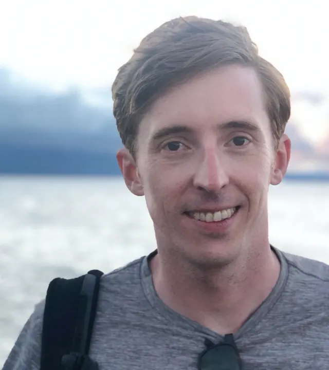 A young man with short brown hair smiles at the camera, standing outdoors near a body of water with a cloudy sky in the background. He is wearing a grey shirt and has sunglasses hanging from his collar.