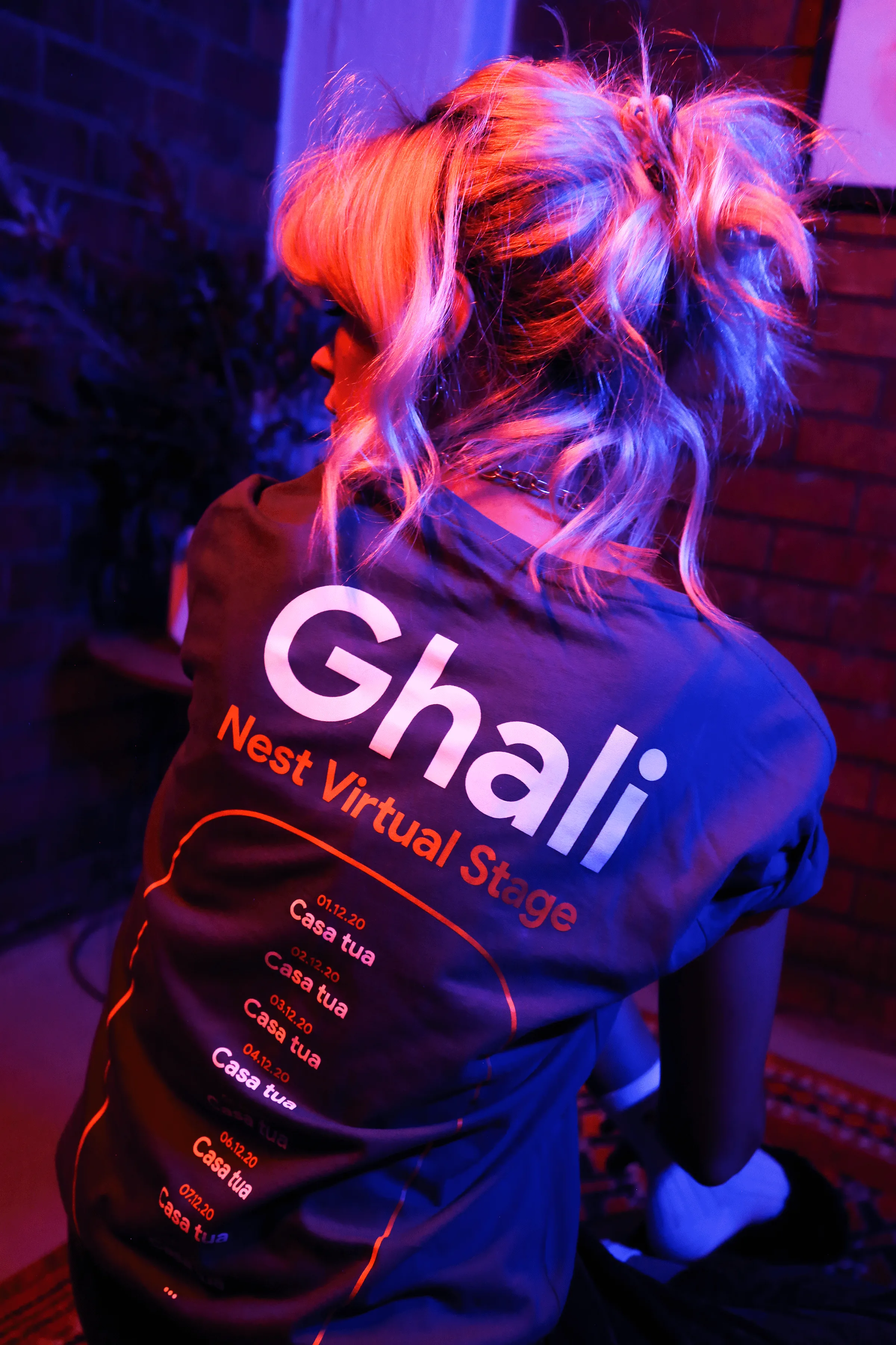 A person with wavy blonde hair in a ponytail is sitting, facing away from the camera. They are wearing a blue shirt with Ghali Nest Virtual Stage and event details printed on the back. The lighting is pink and blue.