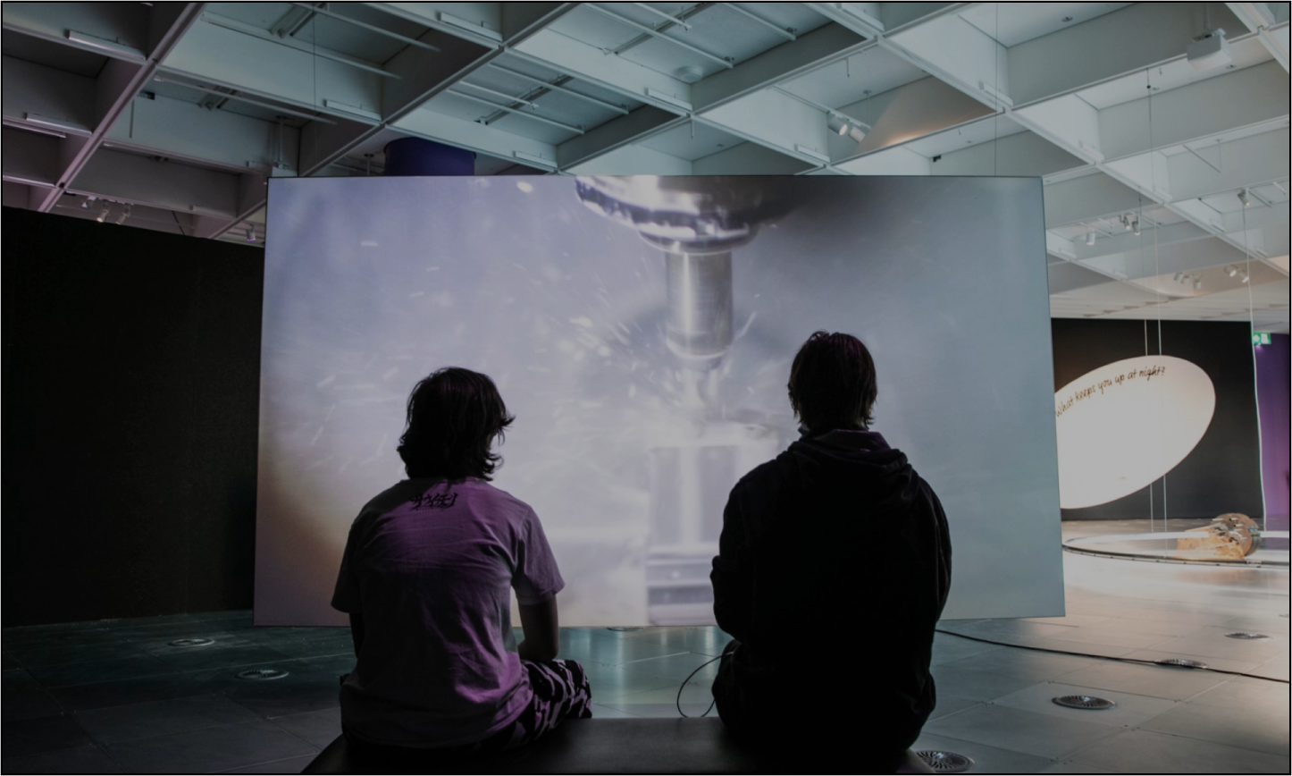 Two people sit on a bench in a dimly lit exhibition space, watching a large screen displaying a close-up of a spinning metallic object, with a spacecraft model visible in the background.