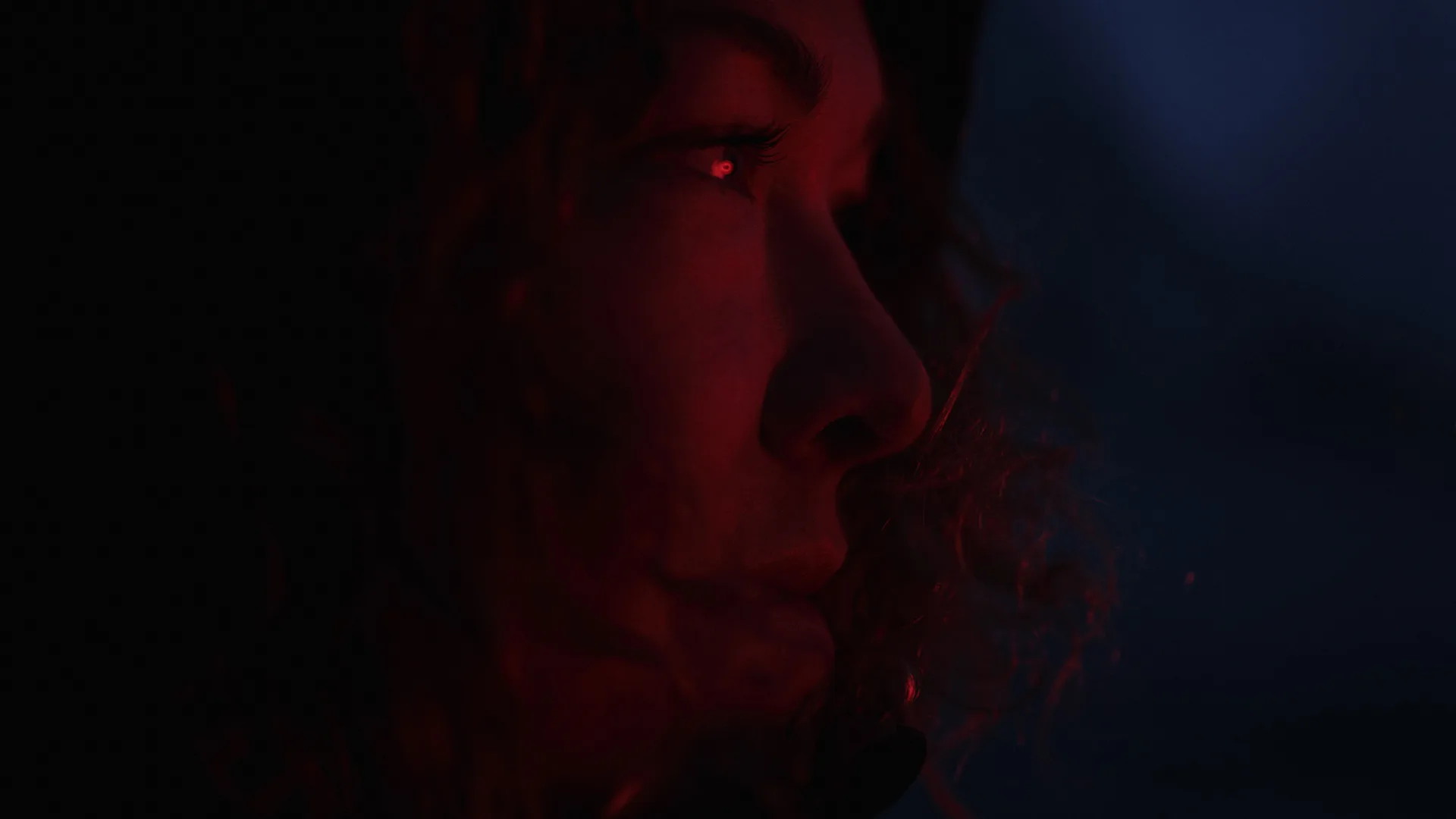 A close-up side profile of a person with curly hair, illuminated by a warm red light against a dark background, highlighting their facial features and contemplative expression.