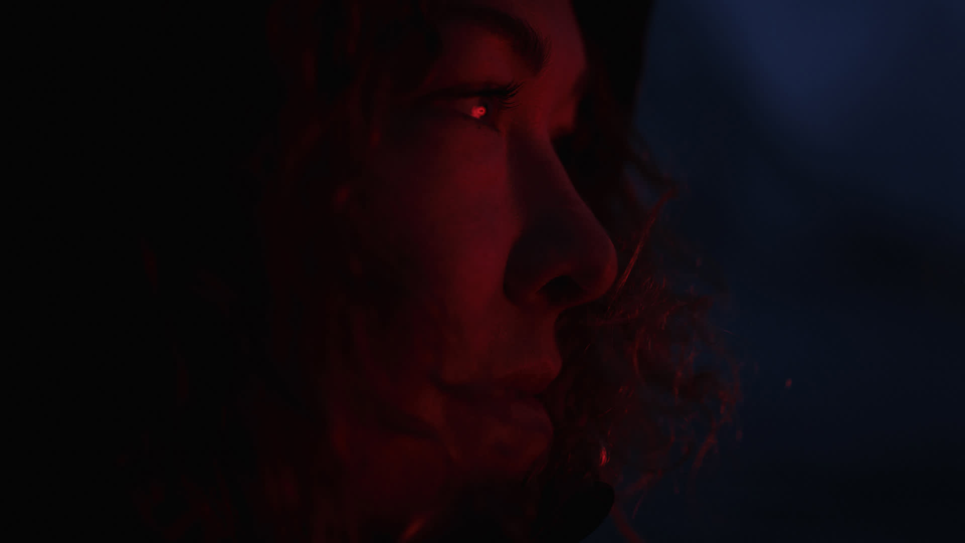 A close-up side profile of a person with curly hair, illuminated by a warm red light against a dark background, highlighting their facial features and contemplative expression.
