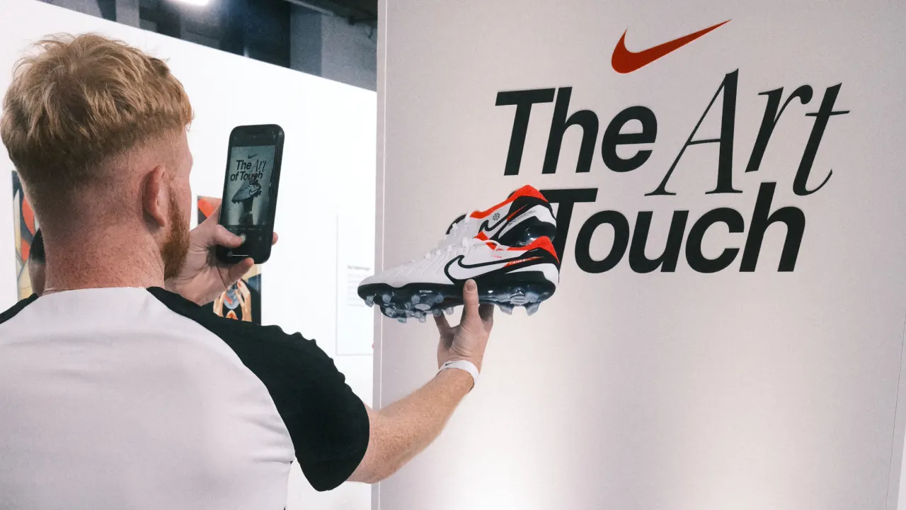 A person holds up a Nike cleat and takes a photo of it with a phone in front of a white wall featuring the Nike logo and the text The Art of Touch.