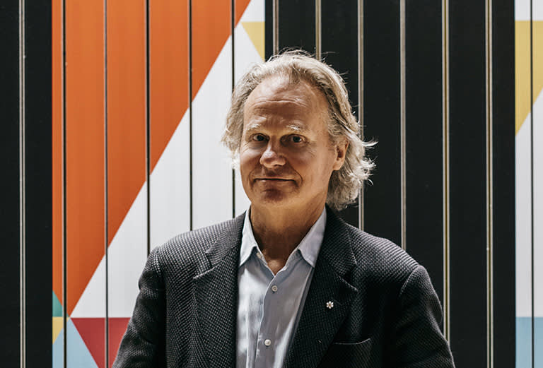 A man with gray hair wearing a dark blazer and light shirt stands in front of a wall with colorful geometric patterns and black vertical lines.