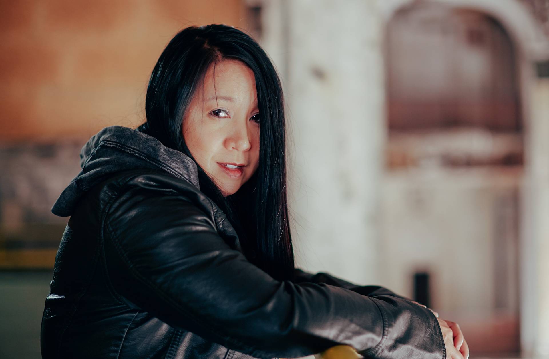 A woman with long black hair and a black leather jacket leans on a yellow railing, looking at the camera. The background is softly blurred, featuring warm tones and a light, textured wall.