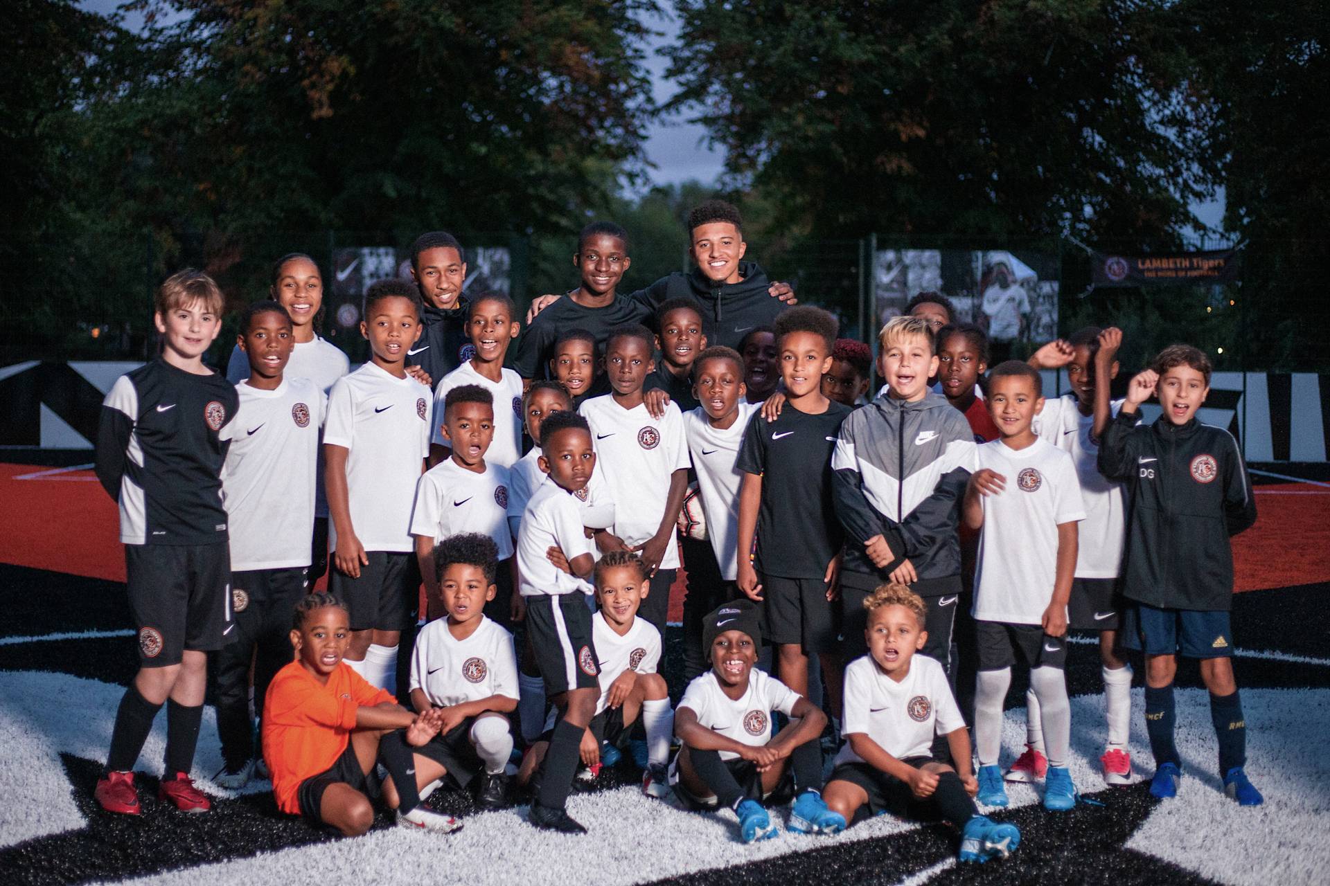 A group of young boys in soccer uniforms pose together for a team photo on an outdoor soccer field, smiling and standing close. Trees and goal posts are visible in the background.