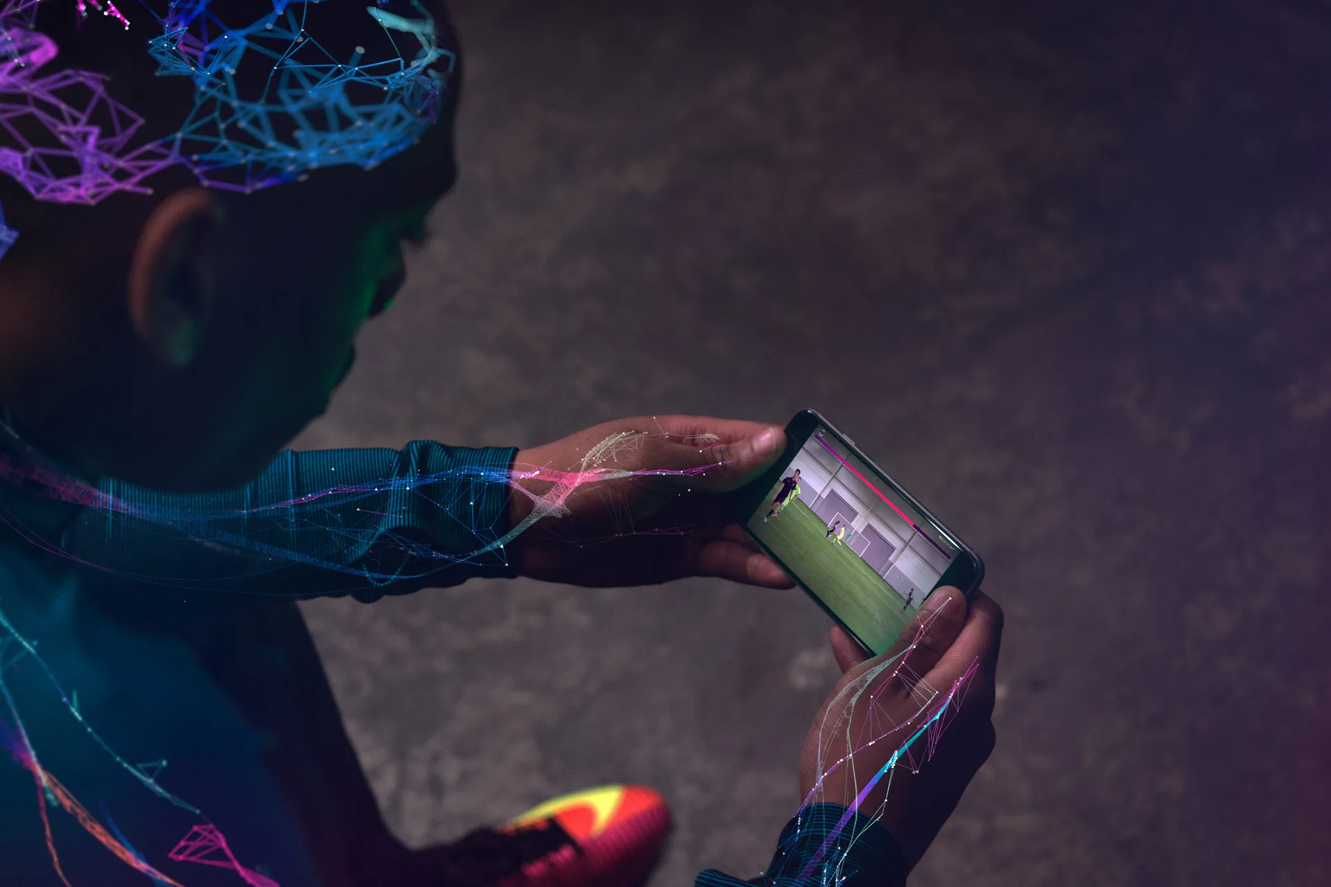 A person wearing athletic gear with digital neon patterns watches a soccer video on a smartphone, viewed from above in a dimly lit environment.
