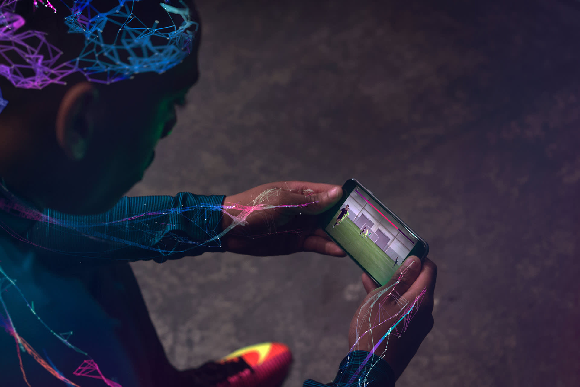 A person wearing athletic gear with digital neon patterns watches a soccer video on a smartphone, viewed from above in a dimly lit environment.