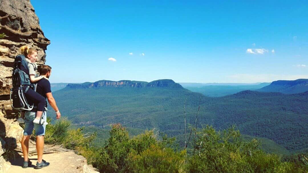 A man carries a child in a backpack carrier while standing on a rocky ledge, overlooking a vast, green valley and distant mountains under a clear blue sky. Dense forest covers the landscape below.