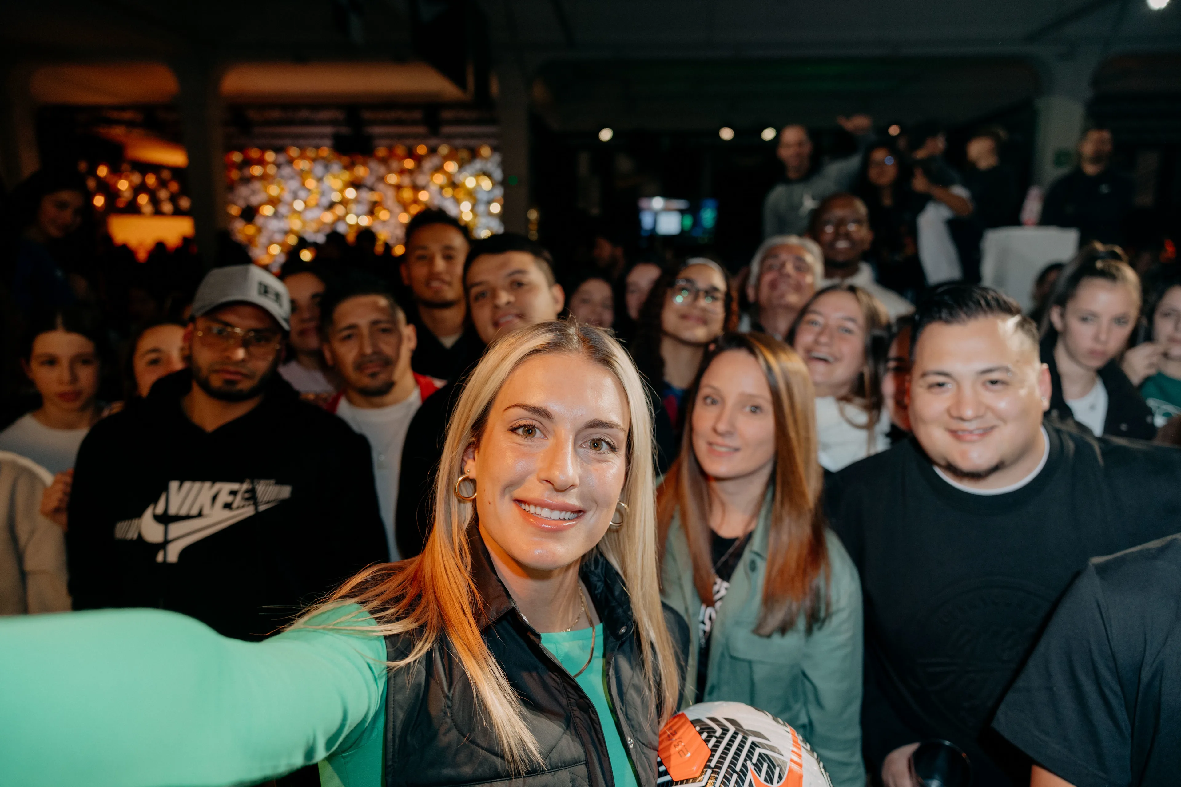 A woman in a green shirt takes a selfie with a large, smiling group of people indoors. She holds a soccer ball and everyone appears happy, posing for the photo in a brightly lit, festive setting.