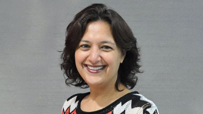 A woman with short, dark, wavy hair smiles at the camera. She is wearing a patterned top with black, white, and red colors. The background is plain and gray.