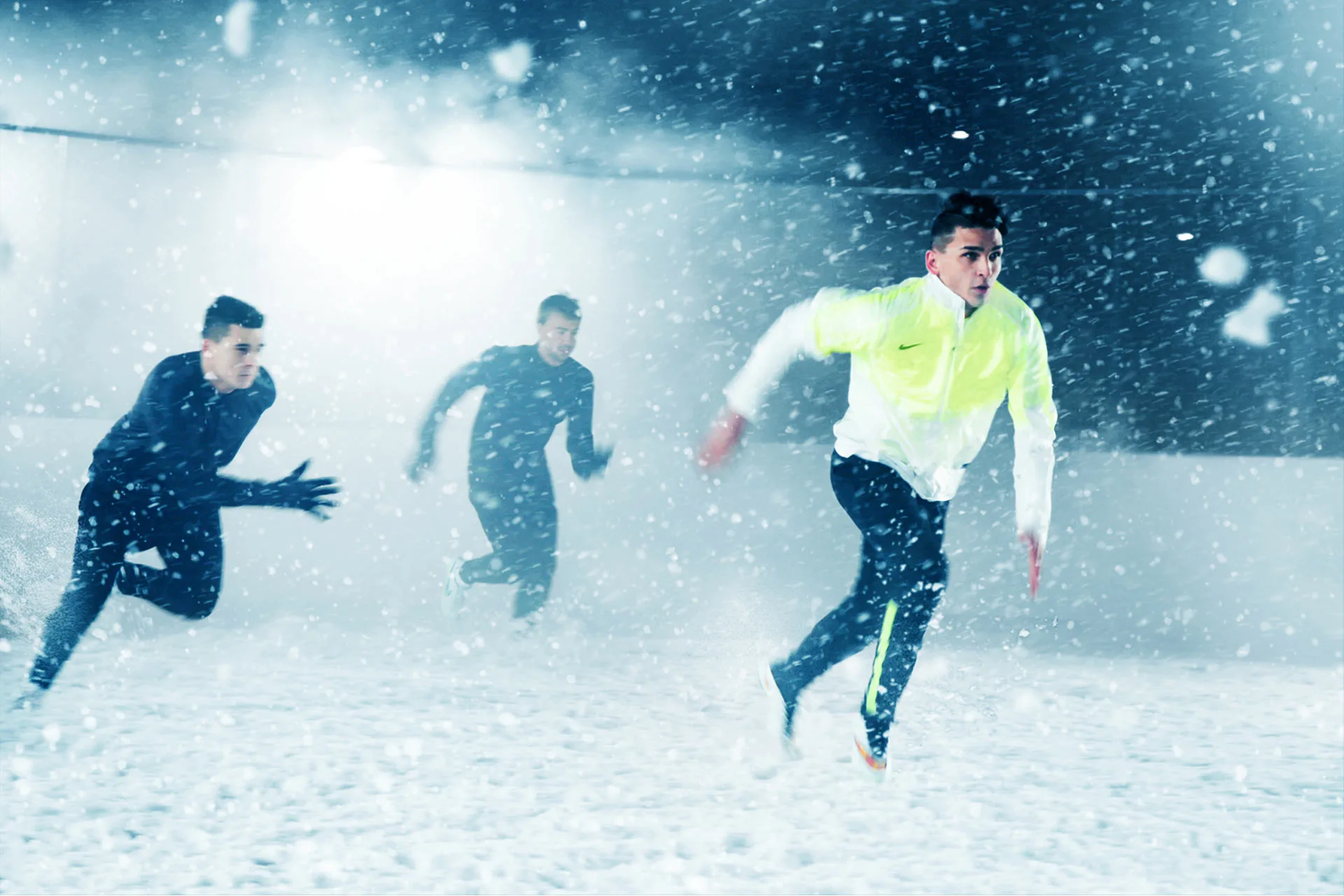 Three people run through heavy snow on a field at night, with one person in a bright yellow jacket leading two others dressed in darker clothing. Snowflakes are falling heavily, and the scene is illuminated by bright lights.
