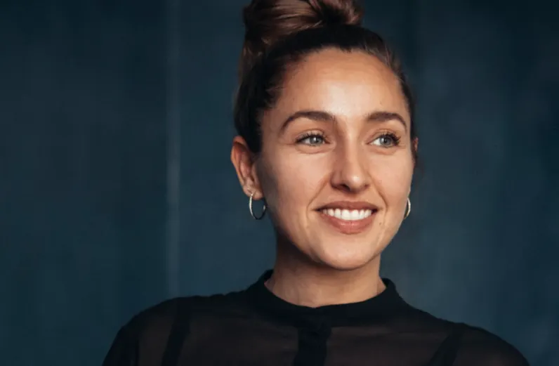 A woman with light skin and brown hair in a top bun, wearing hoop earrings and a sheer black top, smiles while looking slightly to her right against a dark blue background.