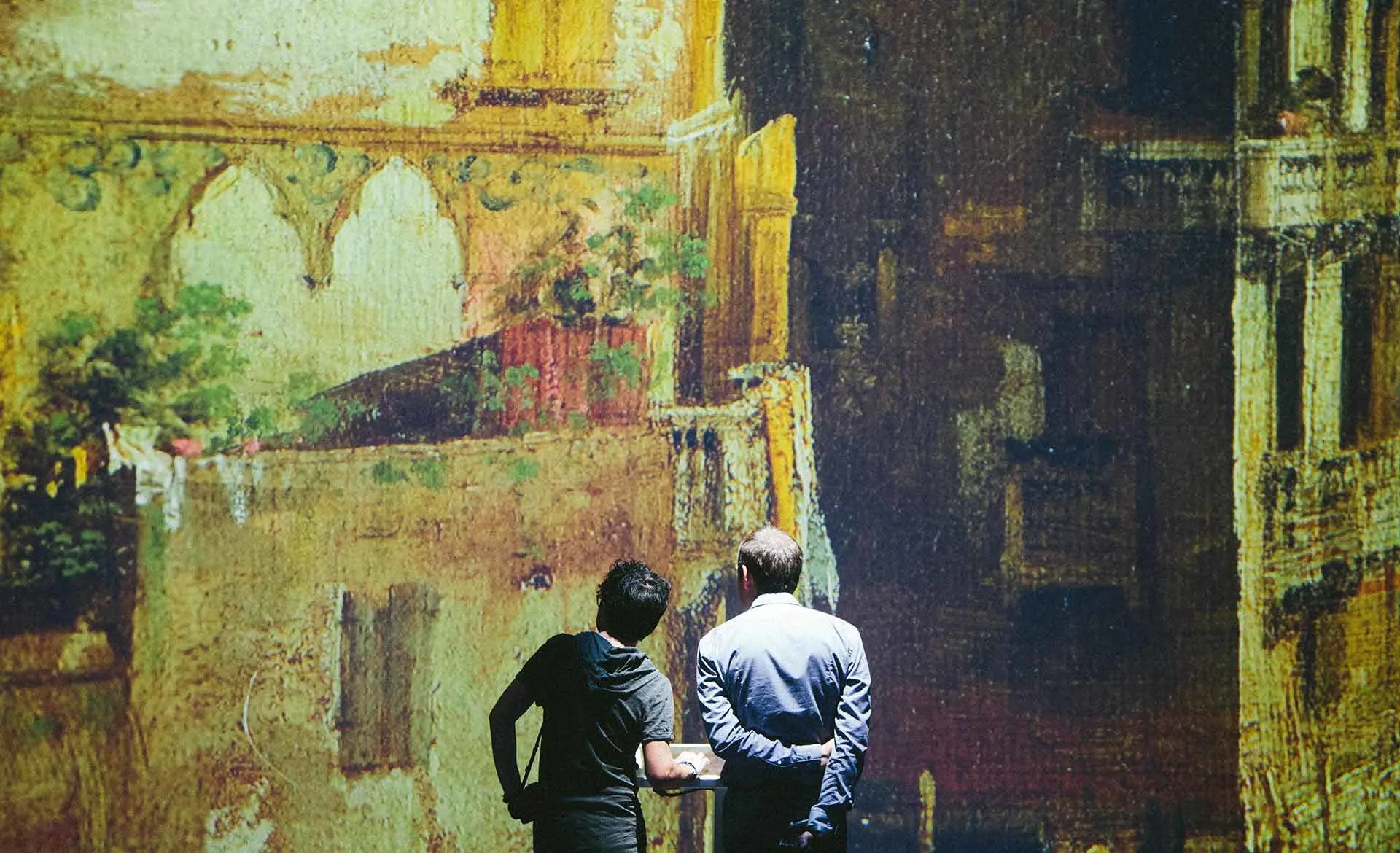 Two people stand closely together, facing a large, illuminated wall displaying a detailed artwork of old, textured buildings with arches and balconies. Their backs are to the camera as they admire the scene.