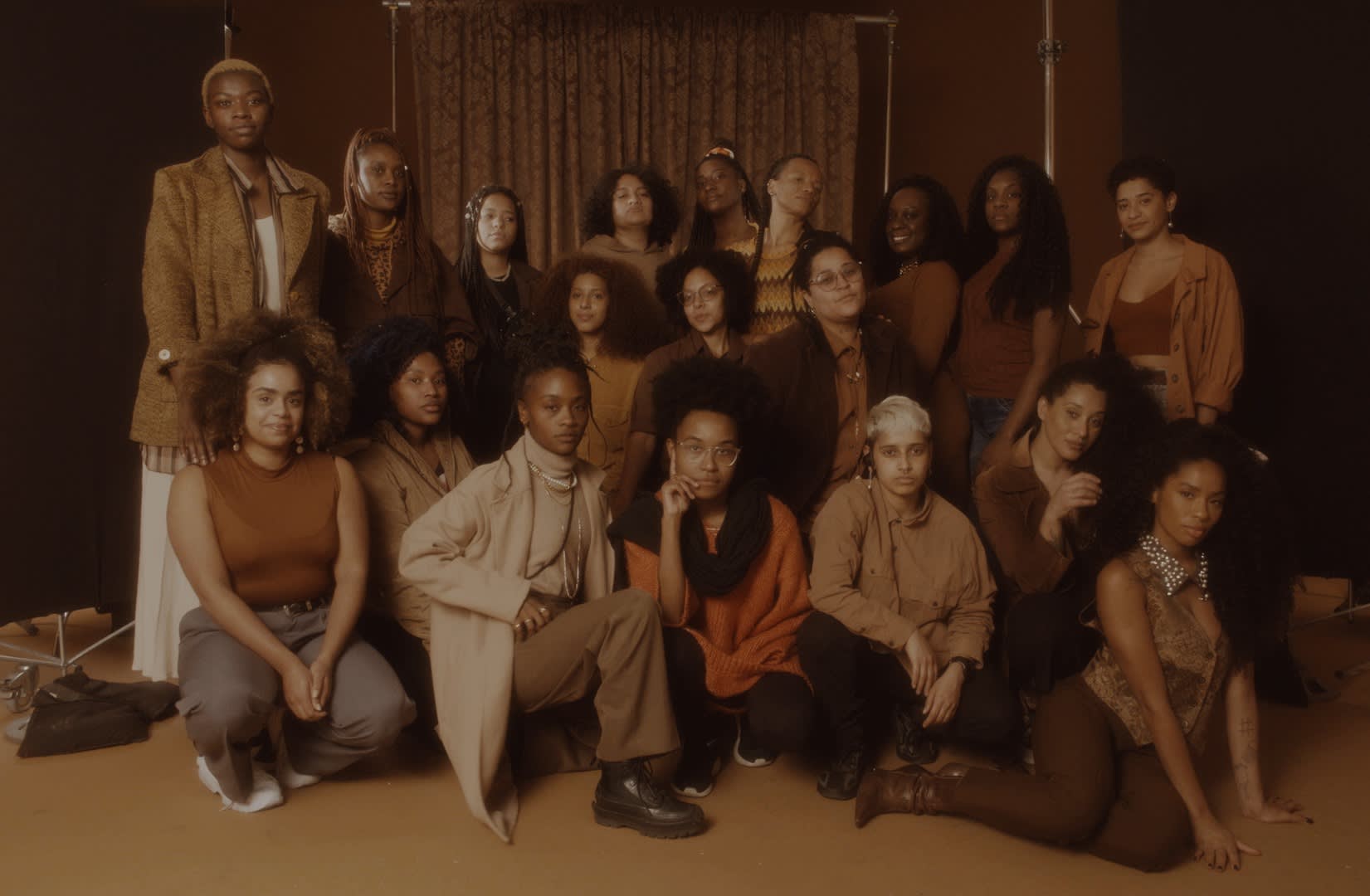 A group of twenty people pose together indoors, wearing earth-toned and brown clothing. Some are standing, while others kneel or sit on the floor. A brown curtain serves as a backdrop. The image has a warm, sepia-toned filter.