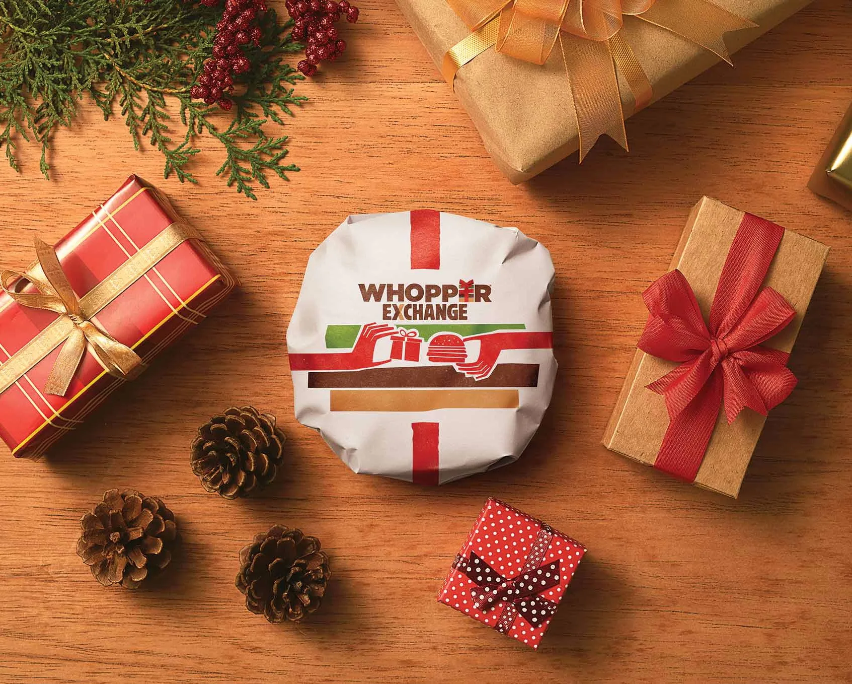 A wrapped Burger King Whopper with WHOPPER EXCHANGE text is surrounded by pinecones, wrapped Christmas gifts, and holiday greenery on a wooden surface.