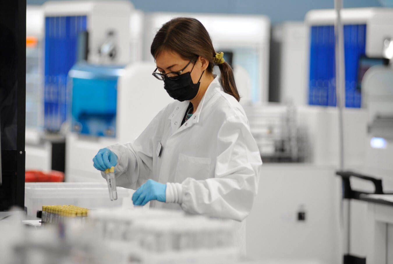 A woman in a white lab coat, black mask, and blue gloves handles test tubes in a laboratory setting filled with scientific equipment and bright lighting.