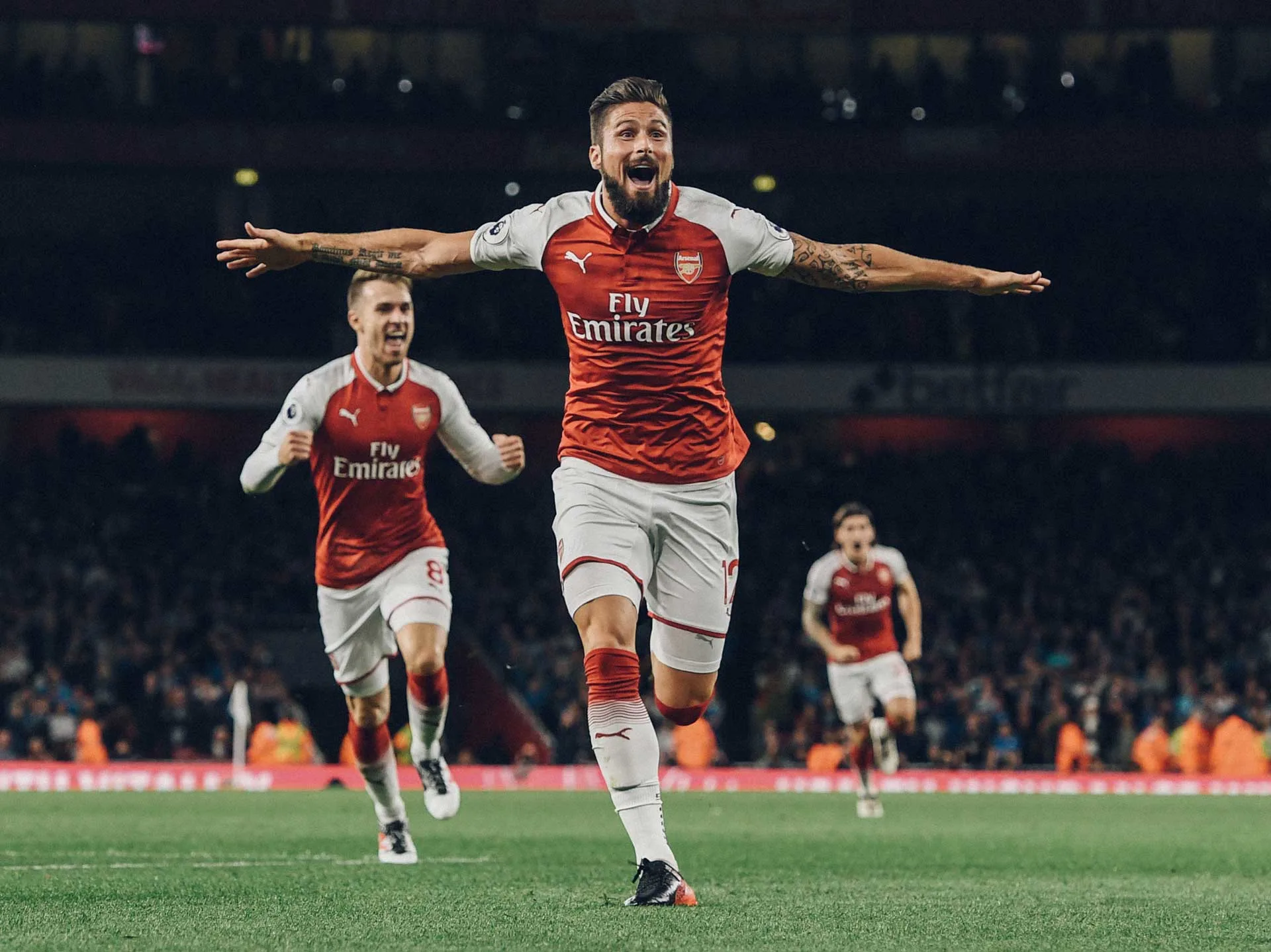 A soccer player in an Arsenal jersey celebrates with arms outstretched after scoring a goal, while teammates run joyfully behind him on a lit stadium field at night.