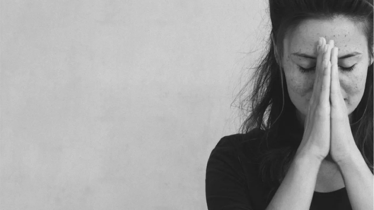 A woman with closed eyes holds her hands together in front of her face in a prayer or meditative gesture, standing against a plain, light-colored background. The image is in black and white.