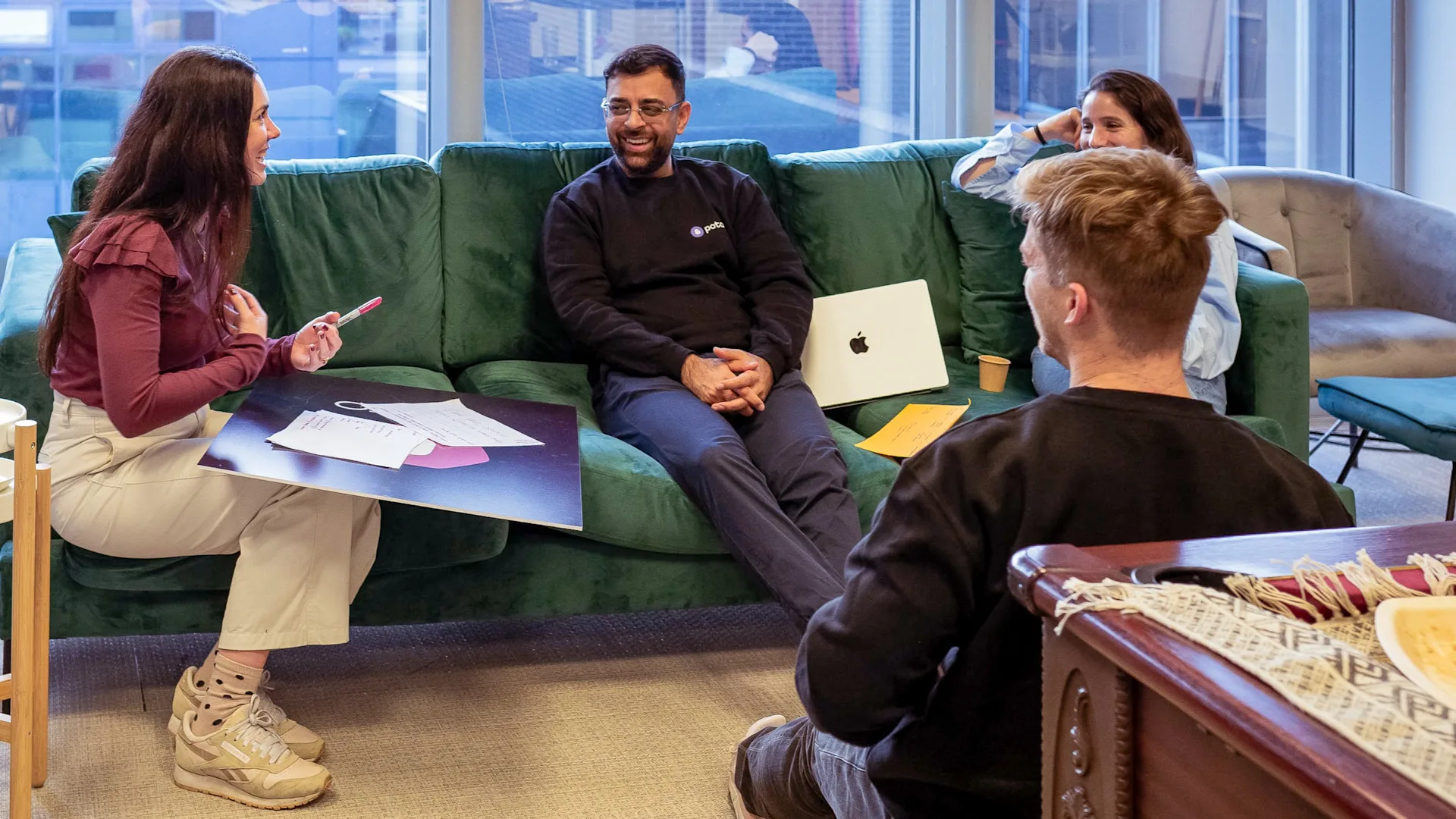 Four people are sitting and talking in a casual office setting. Three are on a green sofa, and one sits on the floor. Laptops, papers, and coffee cups are around them, suggesting a relaxed team meeting.