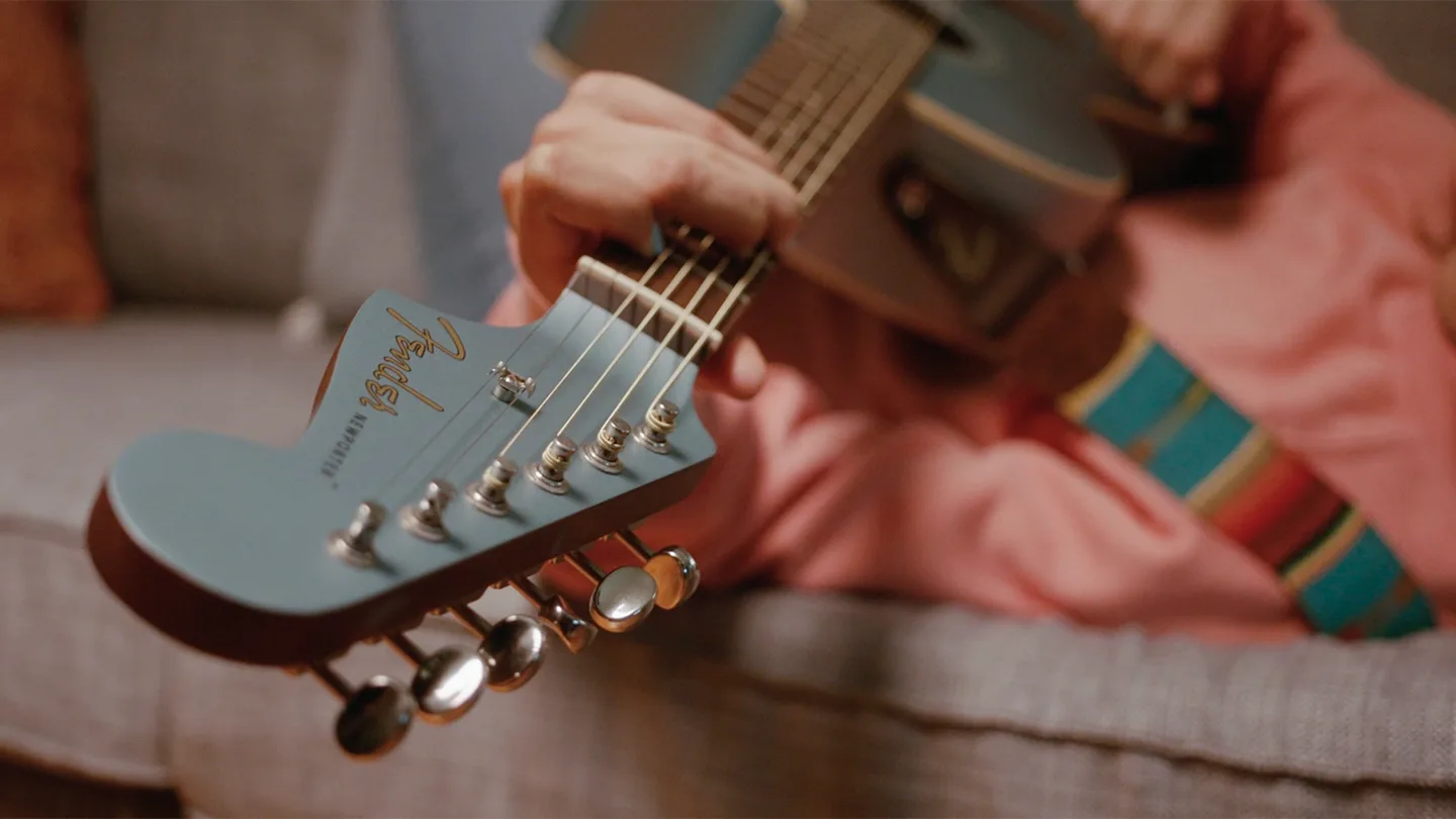 A person in a pink shirt strums a blue Fender acoustic guitar while sitting on a gray couch, with the guitar’s headstock and tuning pegs in focus.