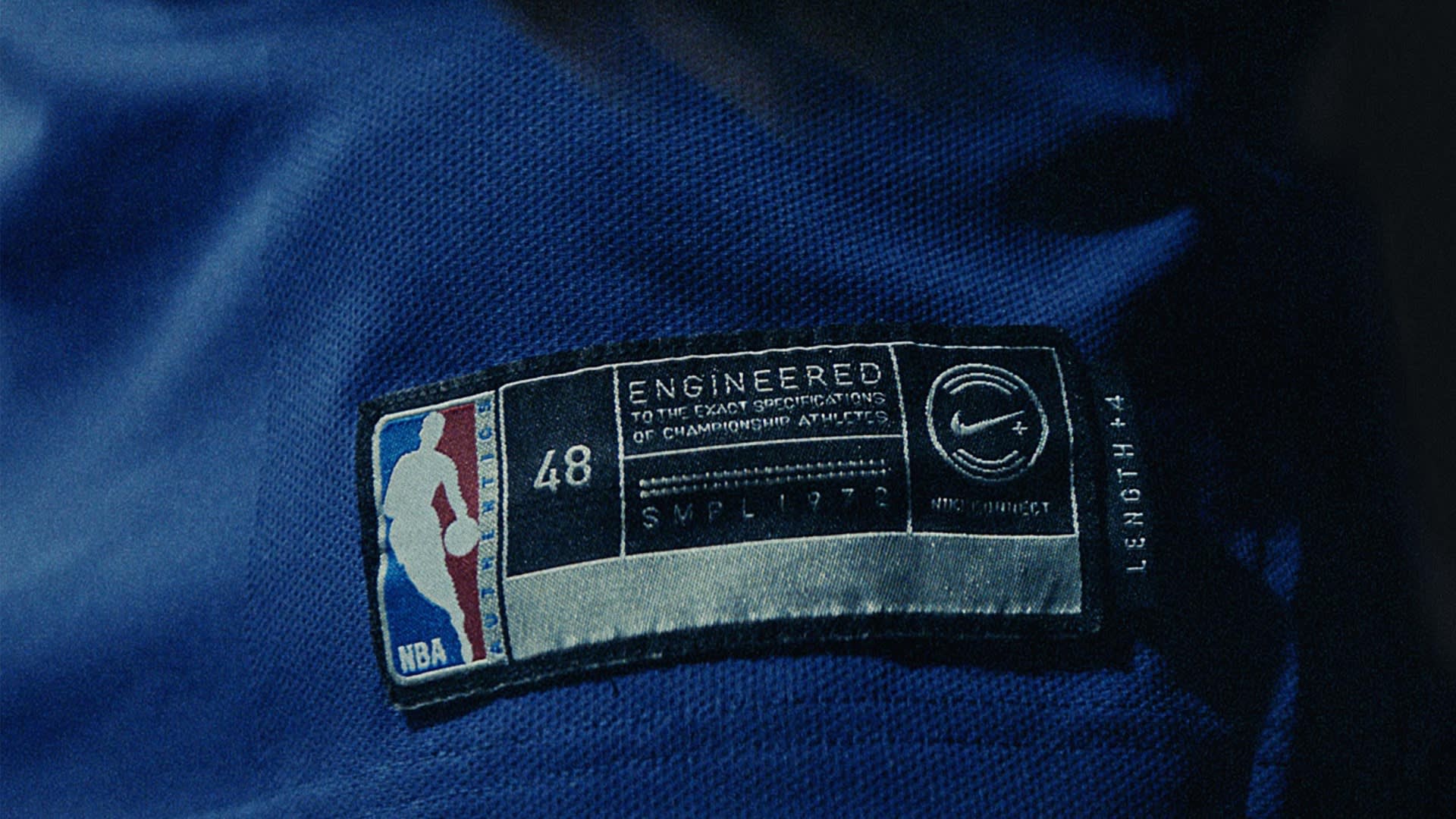 A close-up of a blue NBA jersey features a black and white tag that reads Engineered to the exact specifications of championship athletes, with the NBA logo, size 48, and other details visible.