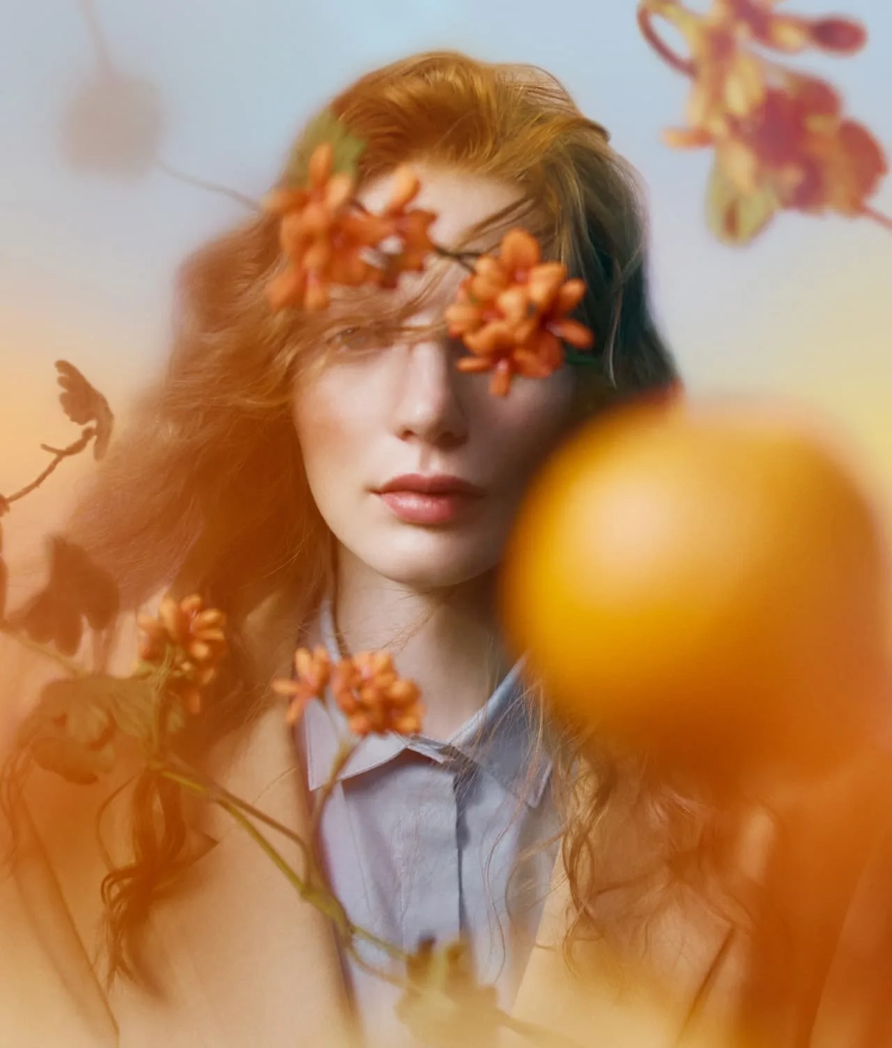 A woman with wavy auburn hair stands behind orange flowers and blurred foliage, softly lit with a dreamy, warm tone. Her face is partially obscured by petals in the foreground.