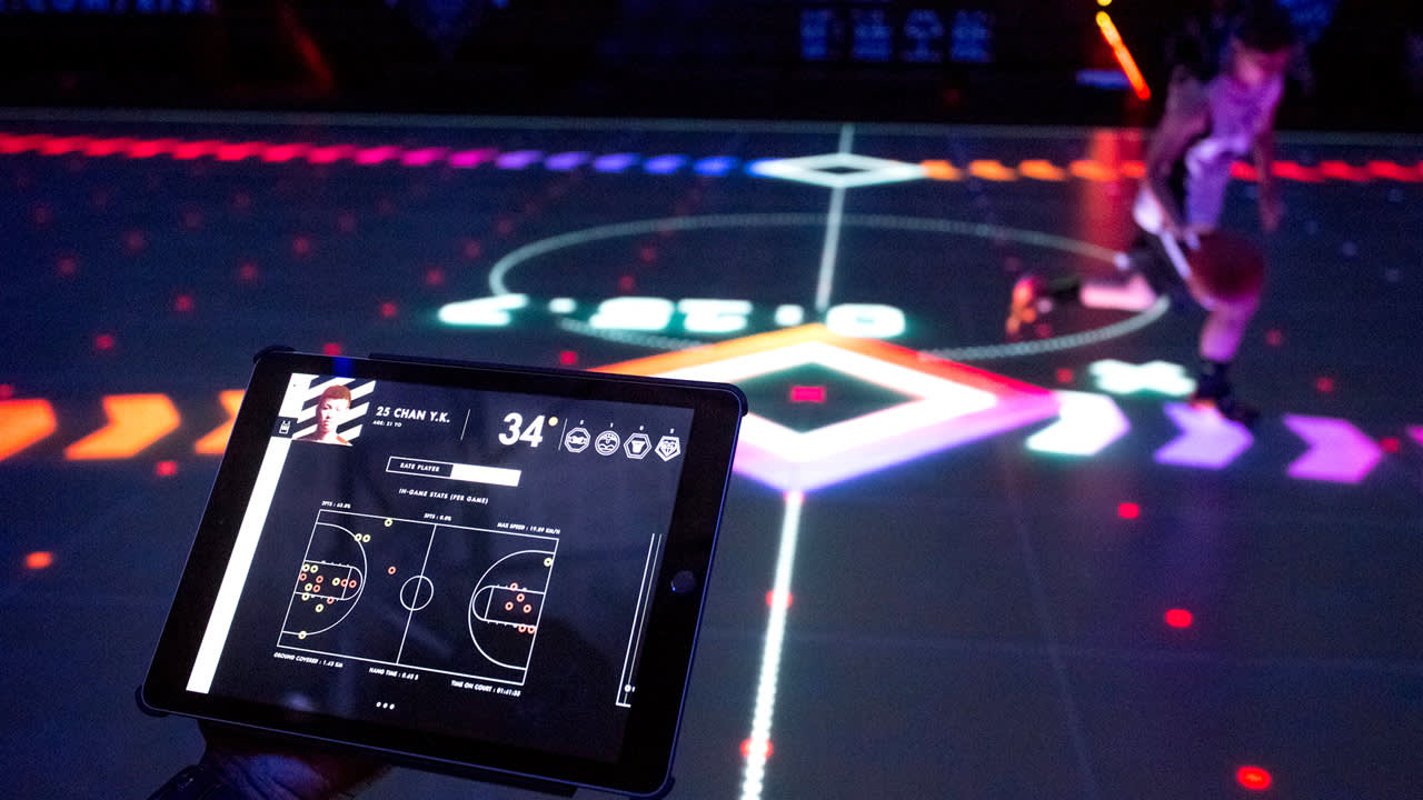A person holds a tablet displaying basketball stats while a player dribbles on an illuminated, high-tech basketball court with vibrant graphics and glowing lines.