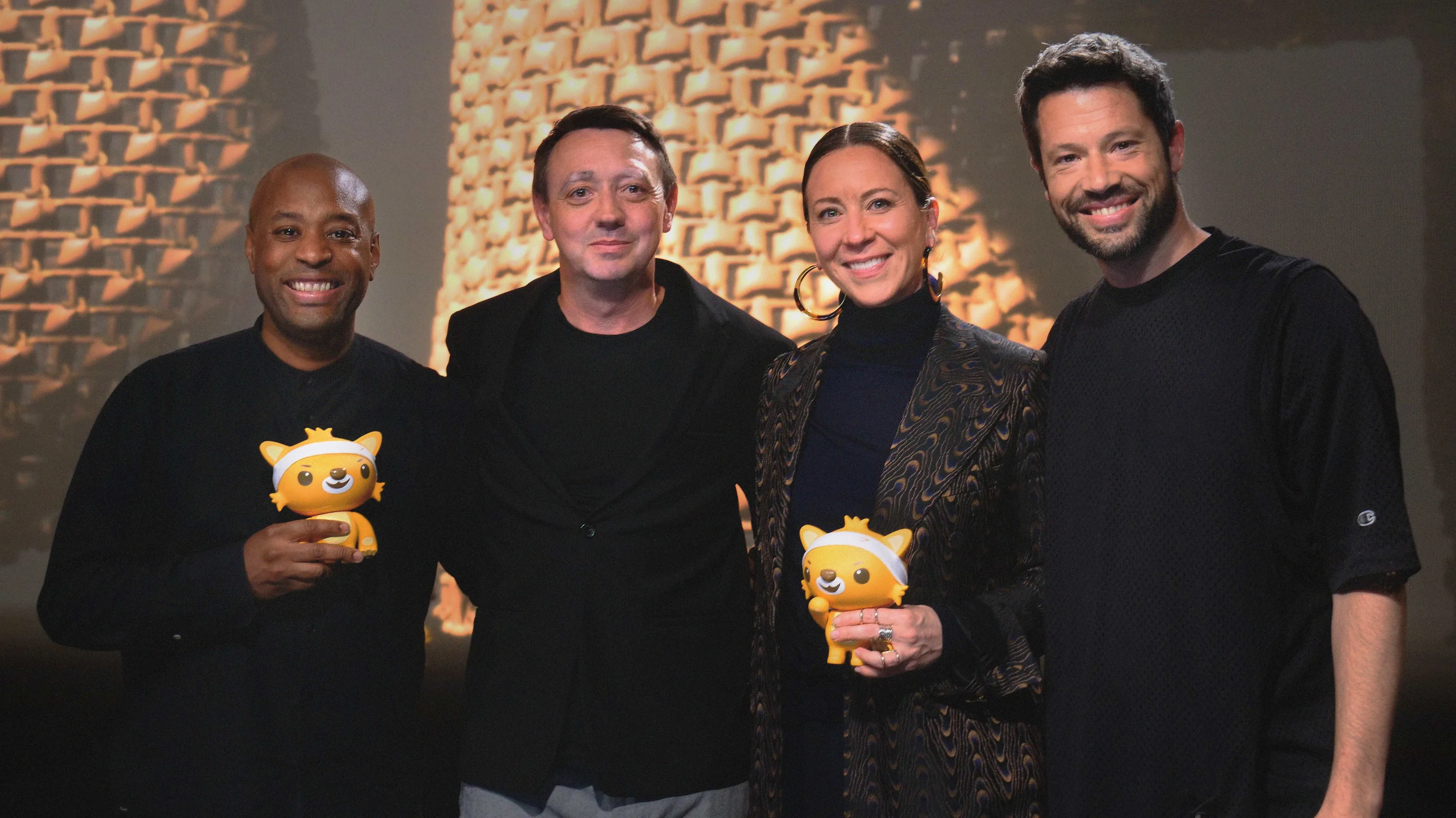 Four people stand together smiling. Two of them hold yellow cat-shaped trophies. The background features a patterned wall with warm lighting.