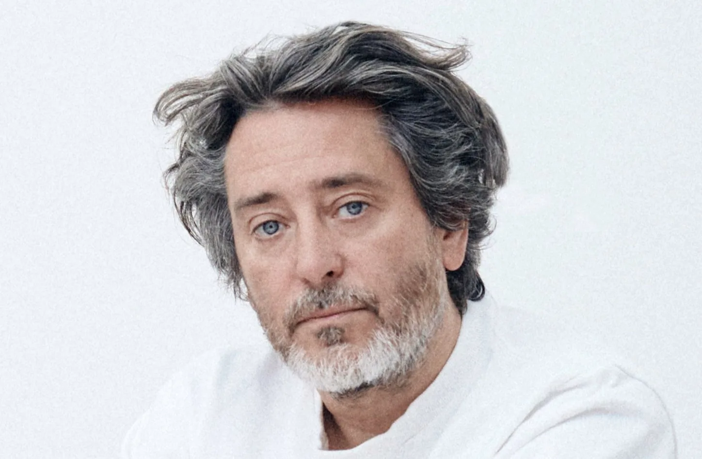 A middle-aged man with wavy gray hair and a short beard looks at the camera with a neutral expression. He is wearing a white shirt and is set against a plain, light-colored background.