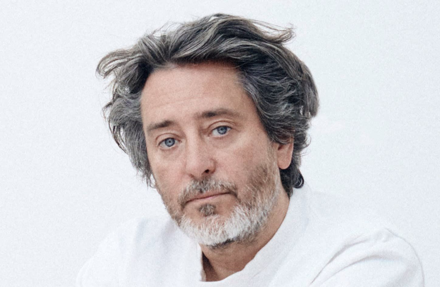 A middle-aged man with wavy gray hair and a short beard looks at the camera with a neutral expression. He is wearing a white shirt and is set against a plain, light-colored background.