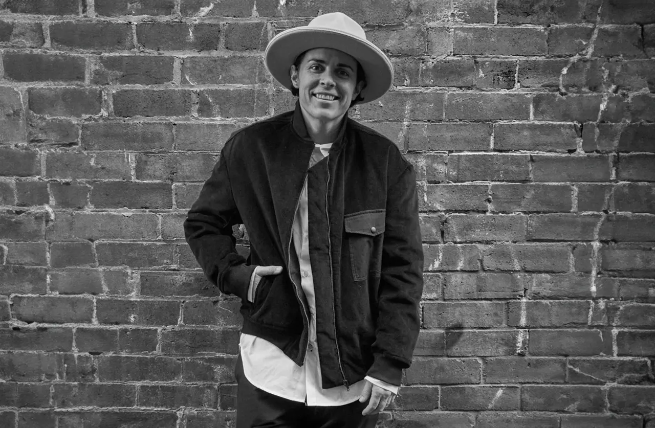 A person wearing a wide-brimmed hat and a jacket smiles while standing in front of a brick wall in a black and white photo.