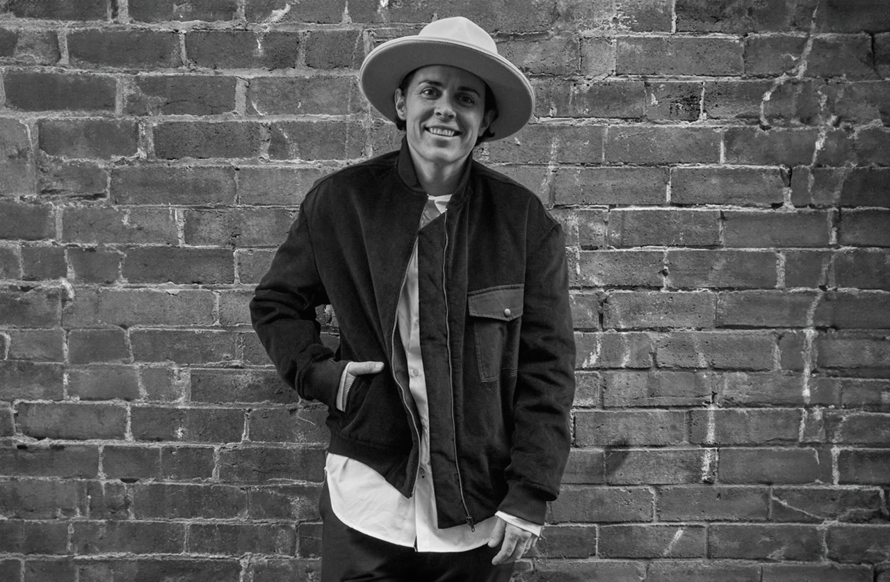 A person wearing a wide-brimmed hat and a jacket smiles while standing in front of a brick wall in a black and white photo.