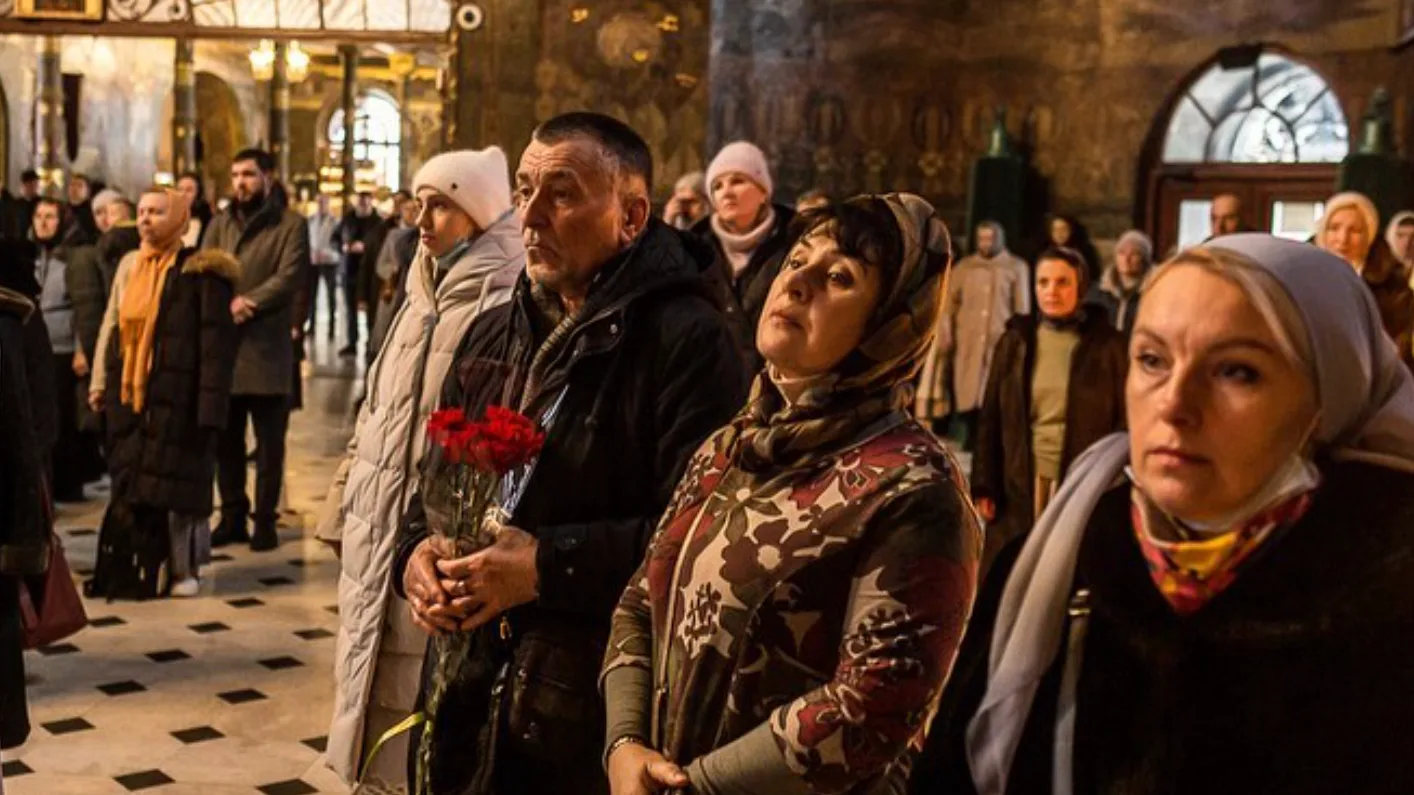 A group of people stand closely together inside a church, some wearing headscarves. One man holds red flowers, and everyone appears solemn, with warm light illuminating the scene.